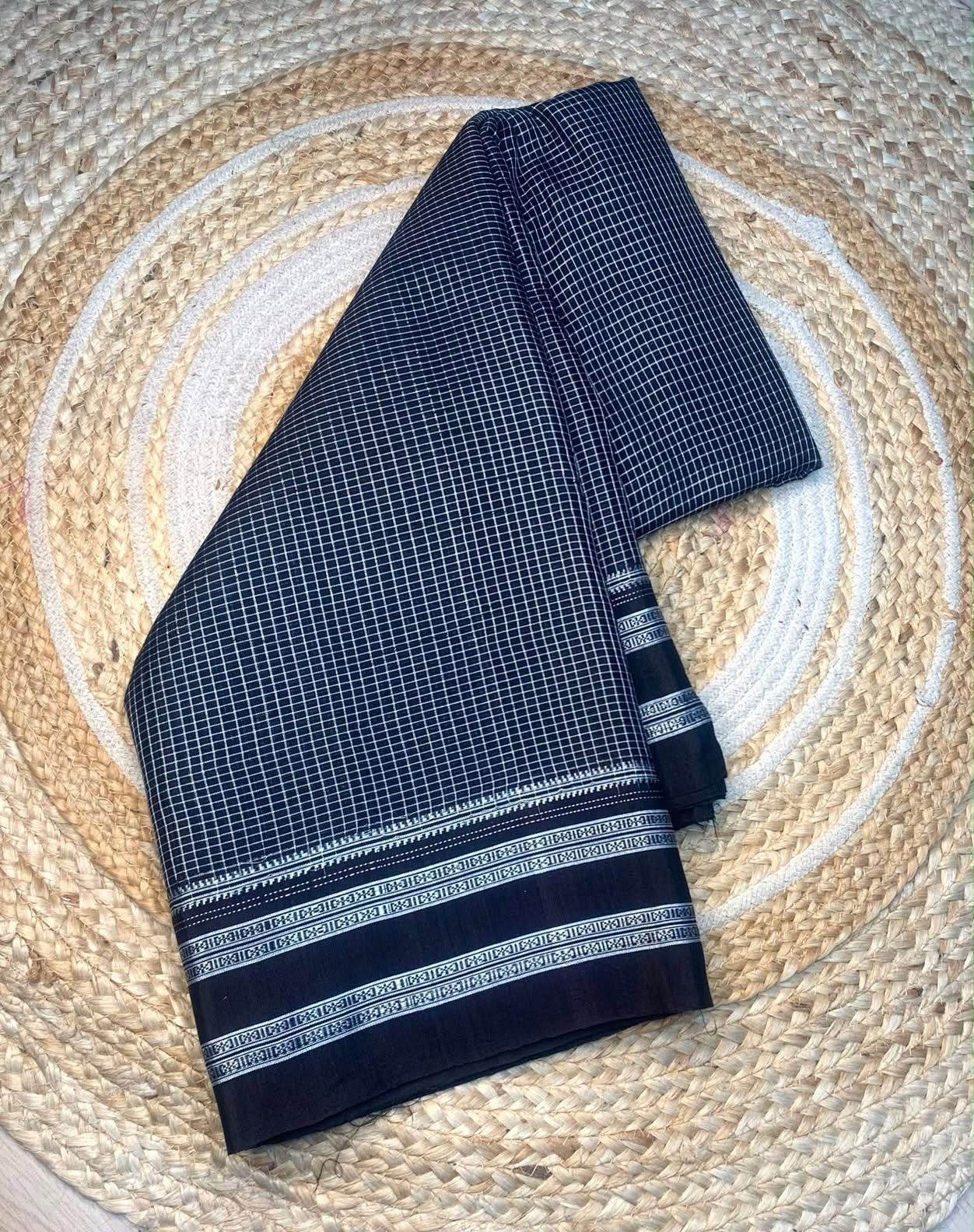 A folded dark navy blue saree with a fine white grid pattern and a decorative border, displayed on a circular woven straw mat.