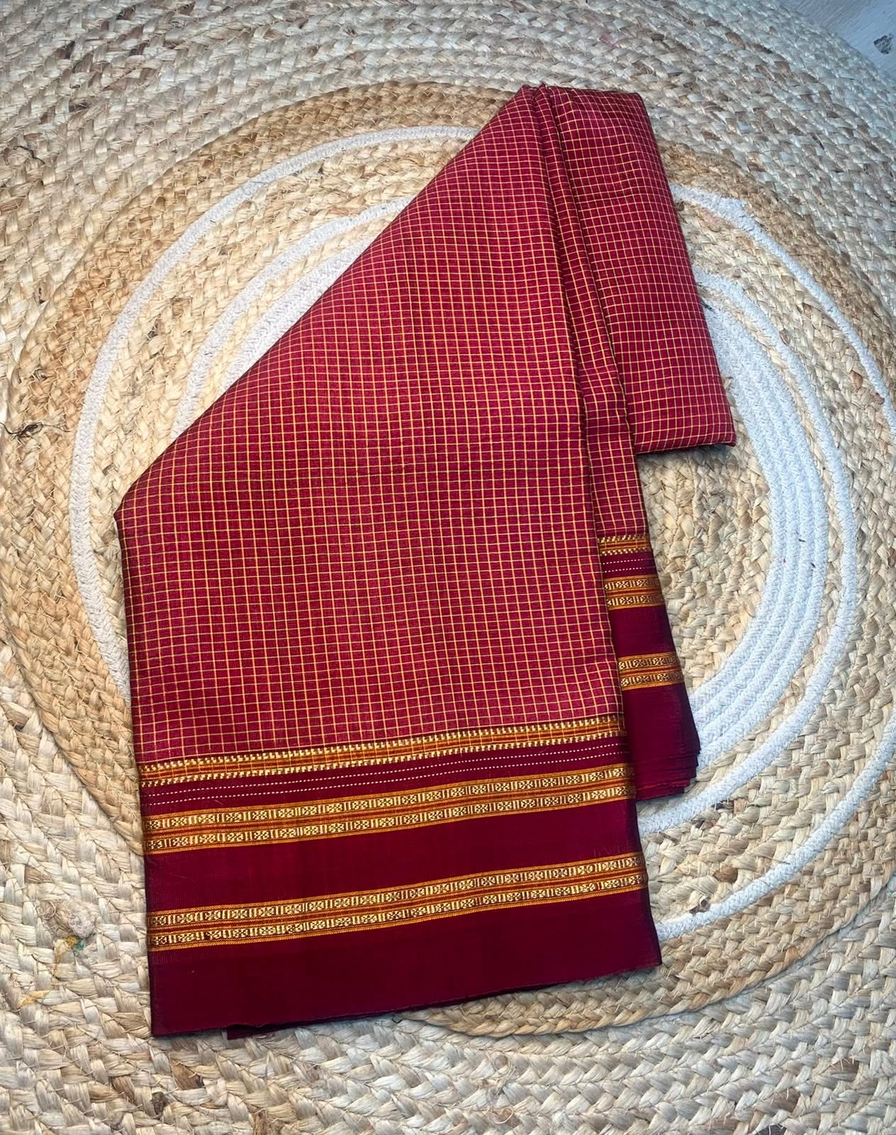A folded red saree with a fine white grid pattern and gold borders, displayed on a circular woven straw mat.
