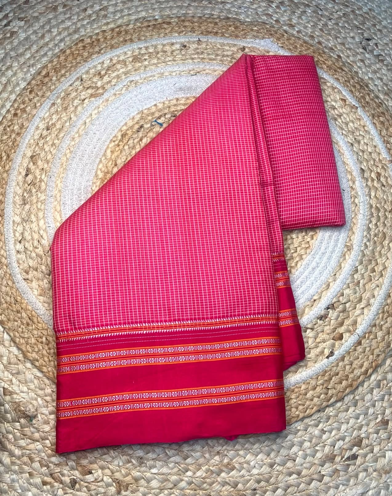 A folded pink and red checkered fabric, likely a saree or sari, with a decorative border, displayed on a woven circular straw mat.