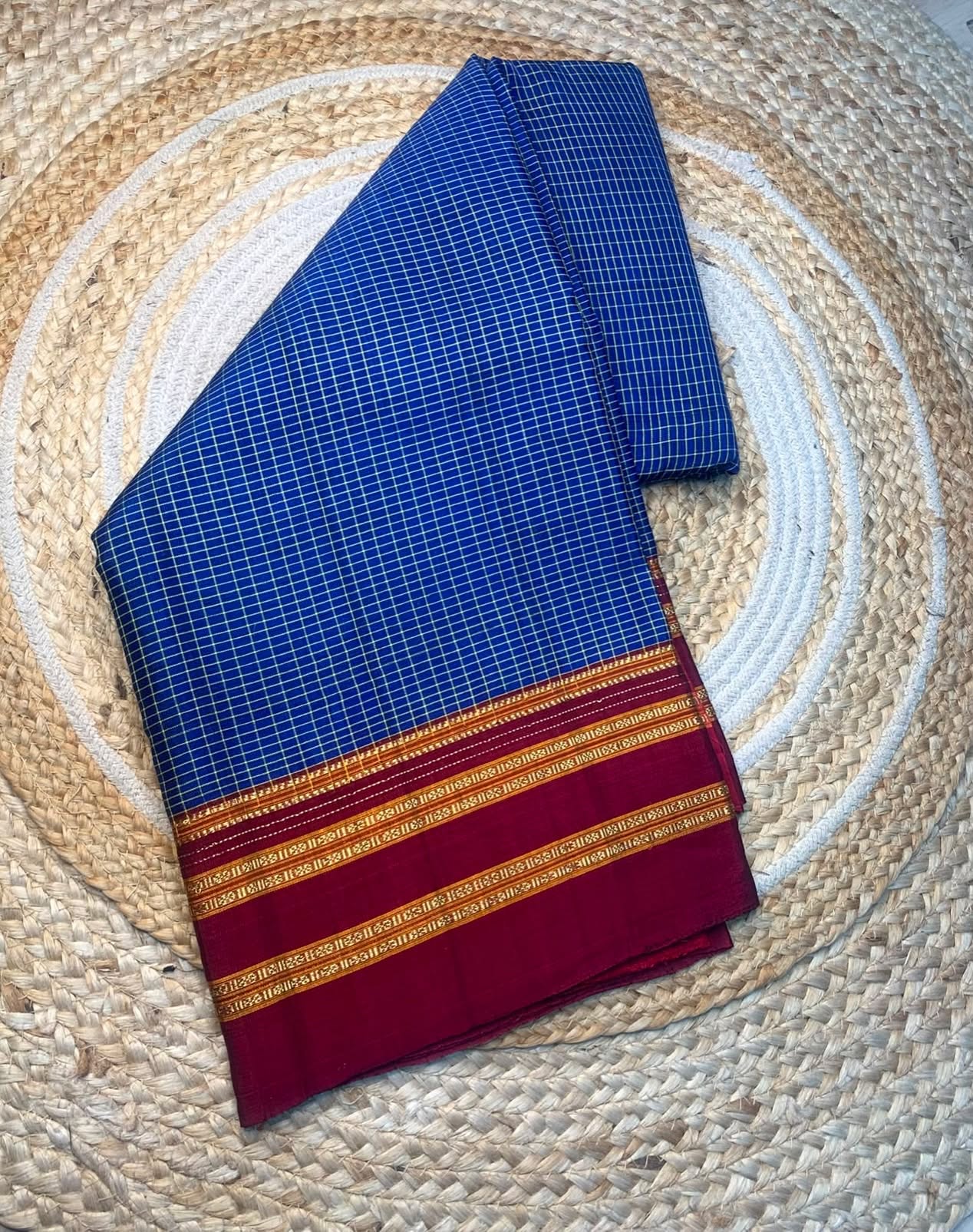 A folded blue and white checkered saree with a deep red border and gold zari stripes, displayed on a circular woven jute mat.