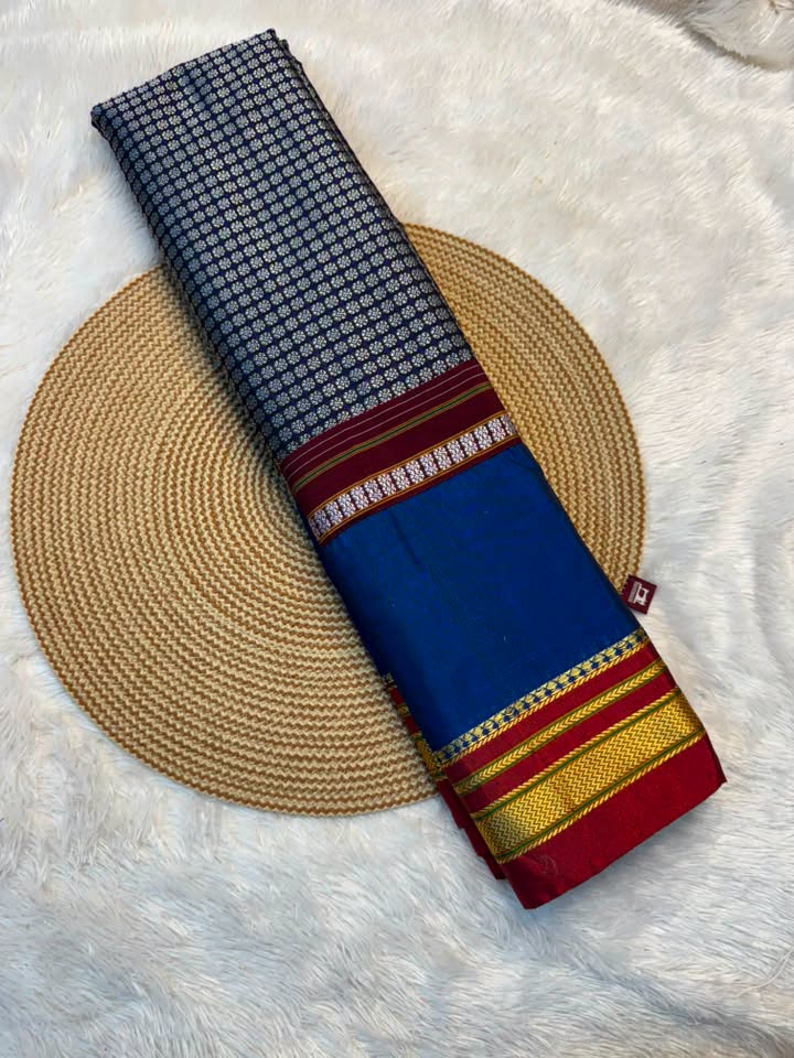 A folded blue saree with a black and white checkered pattern, a royal blue body, and a red and gold border, displayed on a round woven straw mat over a white faux fur background.