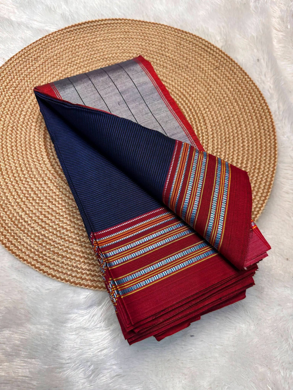 A folded traditional Indian silk saree displayed on a round woven straw placemat over a white faux fur surface. The saree features a navy blue body, a red border, and a silver-gray border with intricate blue, yellow, and orange striped patterns.
