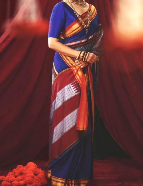 A woman wearing a traditional Indian saree with a royal blue blouse and a maroon, gold, and white patterned drape, posed against a dark red draped backdrop.