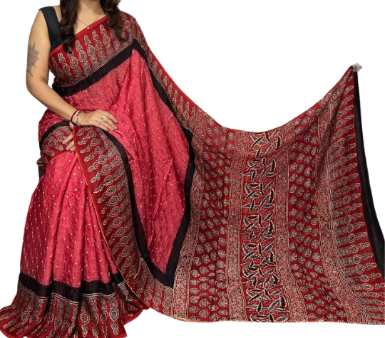 A woman models a red and black patterned saree with a matching blouse against a plain white background. The saree features intricate paisley and floral motifs in red, black, and beige.