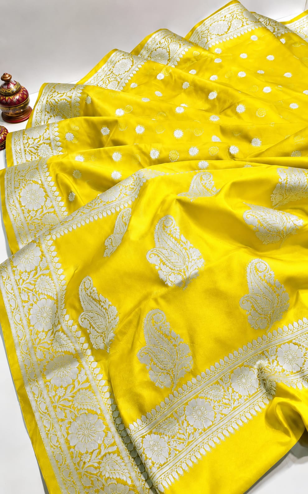 A bright yellow Banarasi silk saree with intricate white zari woven patterns, including paisley motifs and a decorative border, draped elegantly on a white surface.