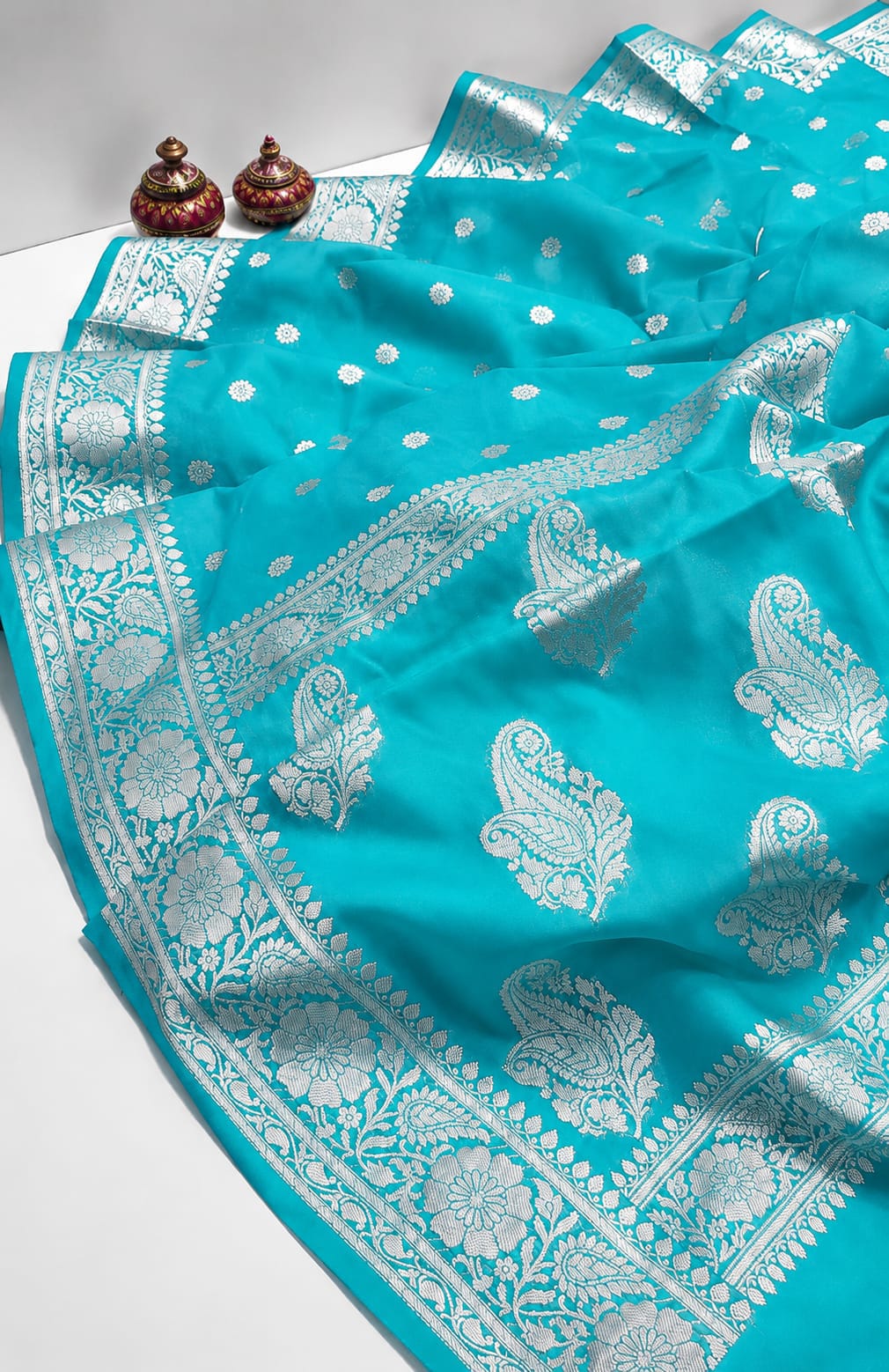 A turquoise blue Banarasi silk saree with intricate silver zari paisley and floral motifs, draped elegantly on a white surface with two small decorative pots in the background.