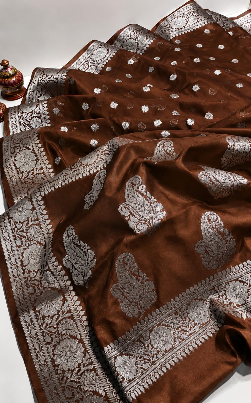 A brown silk saree with intricate silver zari woven paisley and floral patterns, draped elegantly on a white surface.