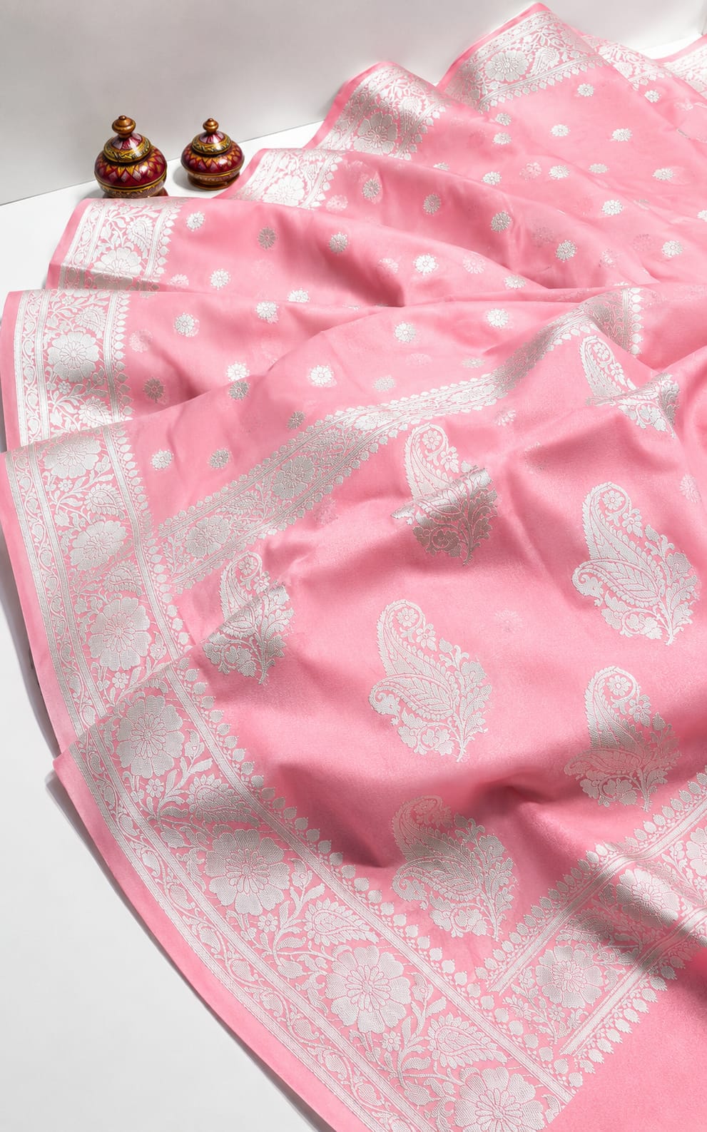 A pink saree with intricate silver paisley and floral motifs, draped elegantly on a white surface with two small decorative pots in the background.
