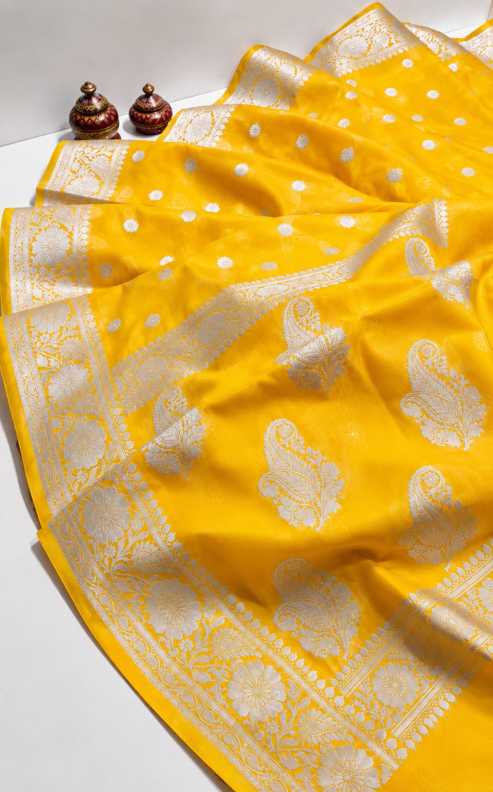 A bright yellow silk saree with intricate silver zari paisley and floral motifs, draped elegantly on a white surface with traditional Indian earrings placed in the corner.
