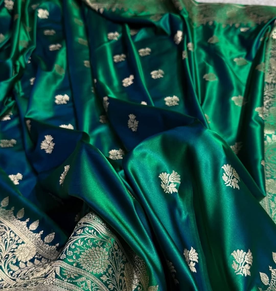 A close-up of a rich emerald green silk fabric, likely a Banarasi saree, featuring metallic silver zari floral motifs and a matching ornate silver border.