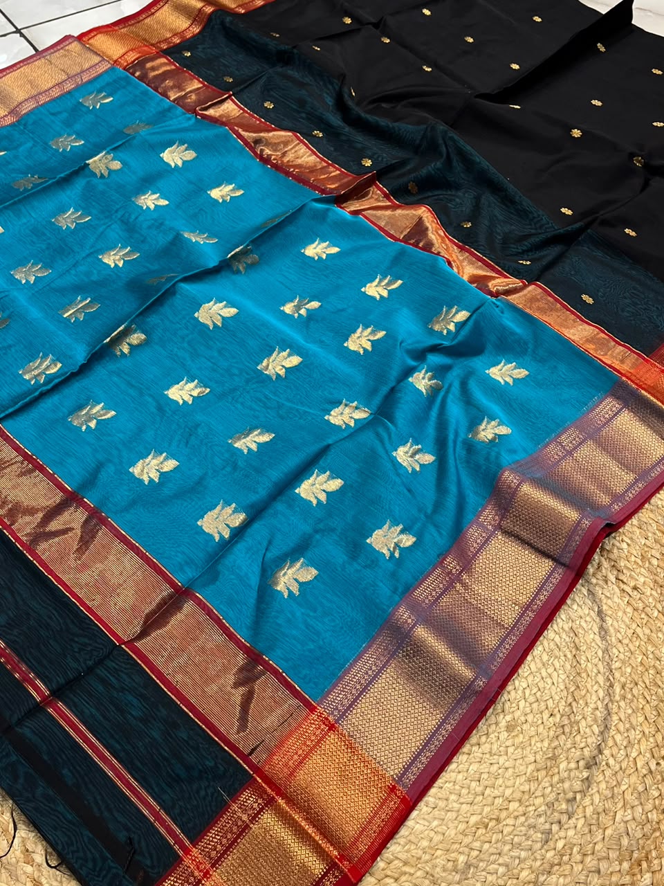 A folded and draped view of a teal blue silk saree with gold floral motifs and a wide gold, red, and navy border, shown alongside a matching black fabric piece with gold dots.