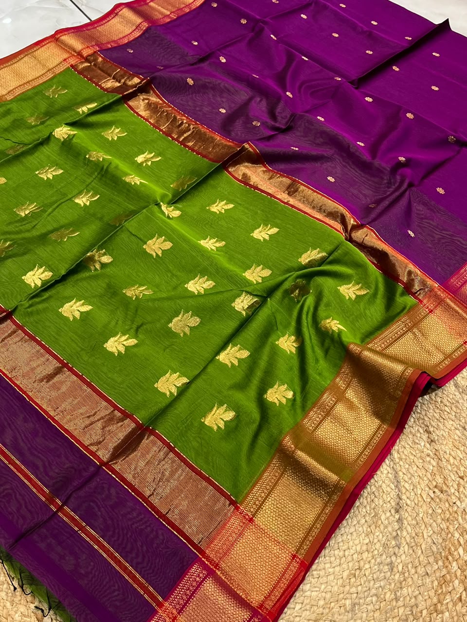 A folded and draped green and purple silk saree with gold zari floral motifs and a wide gold and red border, laid out on a textured beige surface.