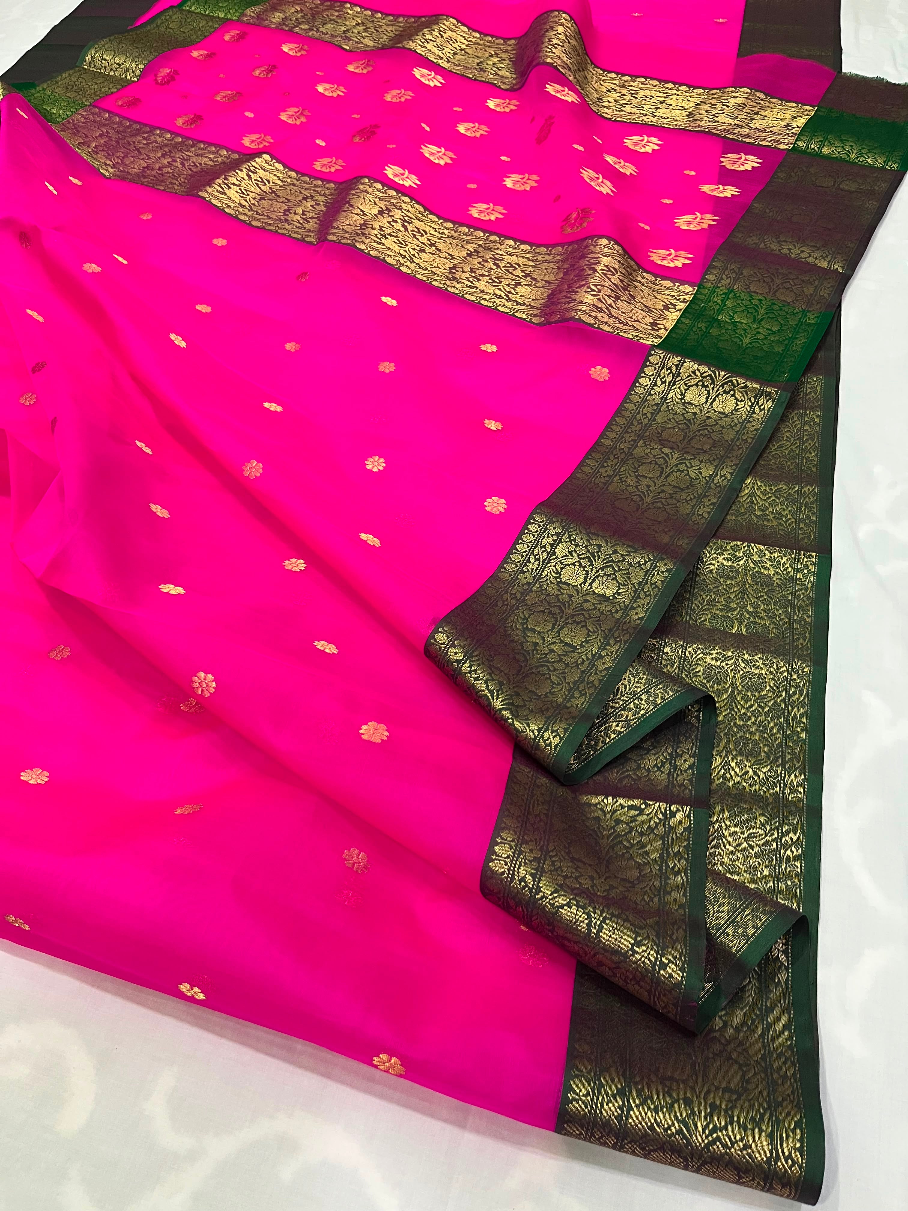 A bright pink silk saree with scattered small gold floral motifs, featuring a wide dark green and gold zari border and a matching pink blouse piece draped on top.