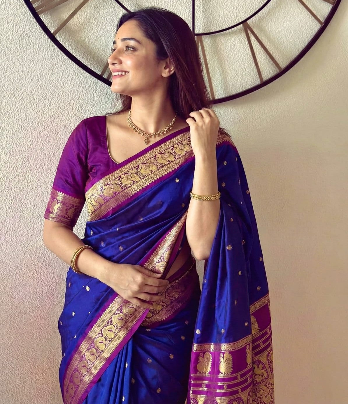 A woman wearing a royal blue and purple silk saree with ornate gold zari borders, standing against a beige wall with a decorative circular clock behind her.