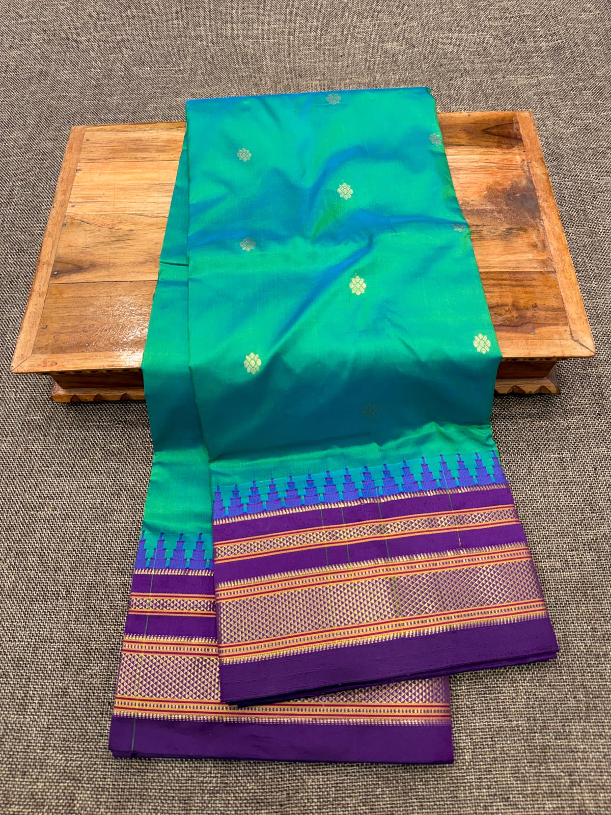 A green and purple silk saree with gold zari patterns, draped over a wooden tray on a gray textured carpet.