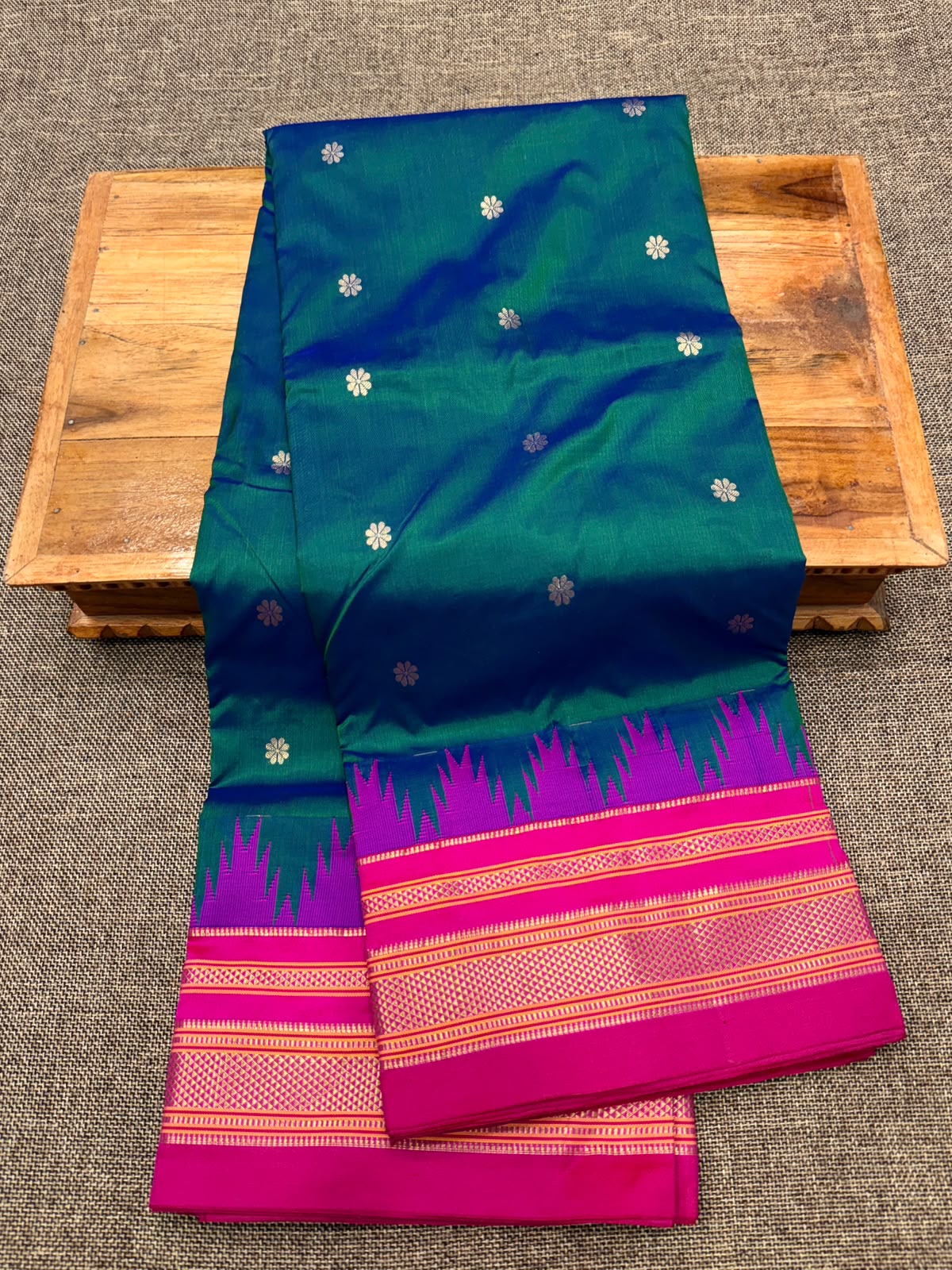 A folded green and magenta pure silk ilkal saree with gold zari motifs, displayed on a wooden tray over a gray textured background.