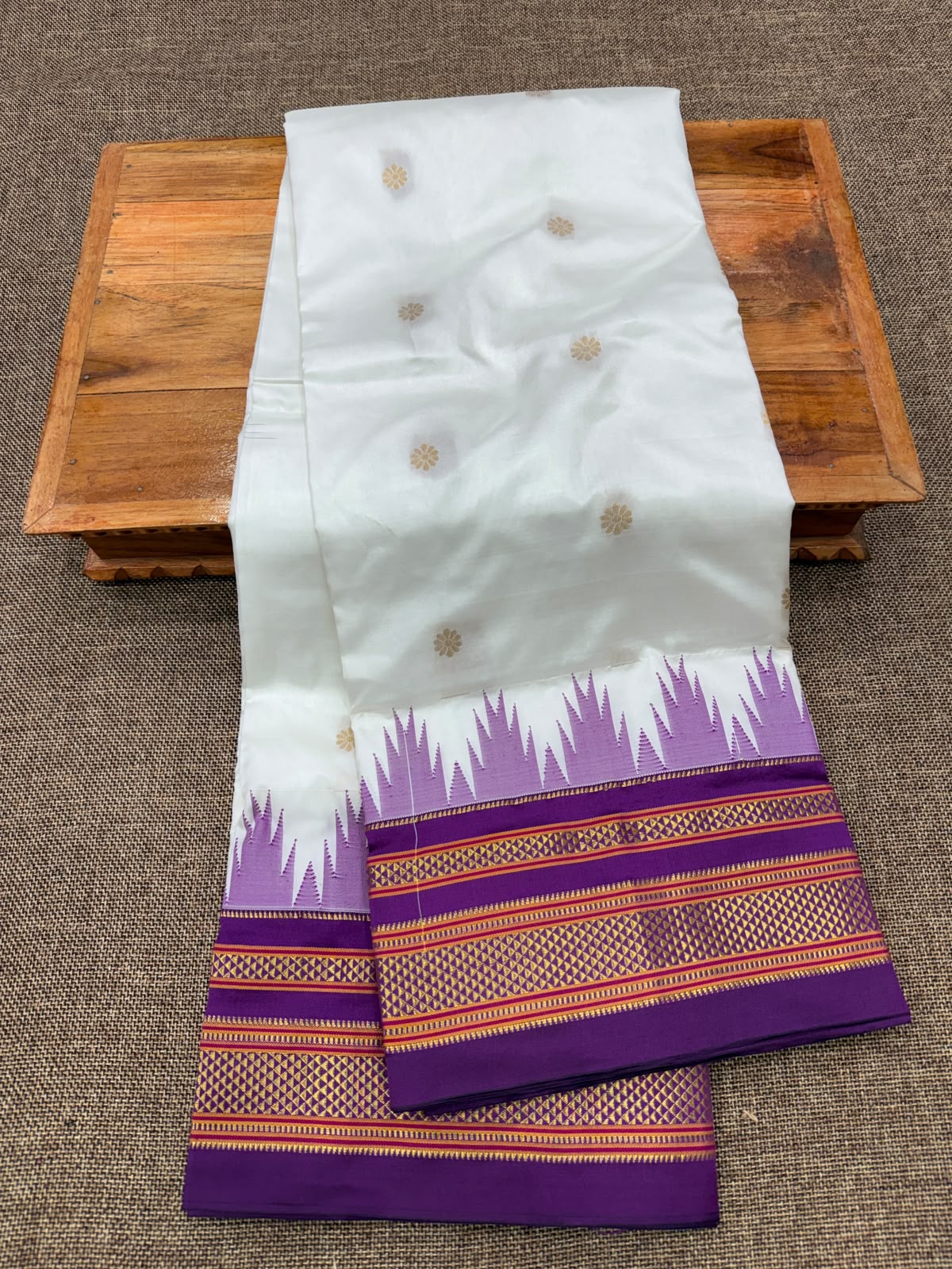 A white silk saree with gold zari motifs and a purple border, draped over a wooden tray on a textured gray surface.