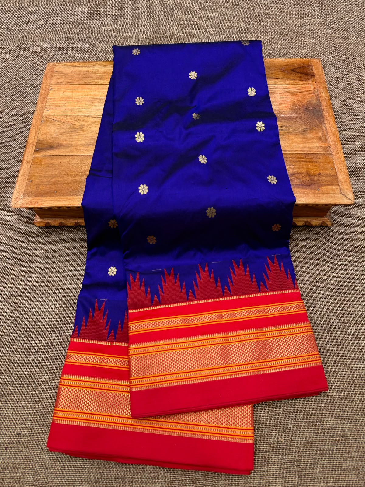 A royal blue and red woven fabric, likely a saree, draped over a wooden table on a textured gray carpet. The fabric features small gold floral motifs on the blue section and a wide red border with gold zari patterns.