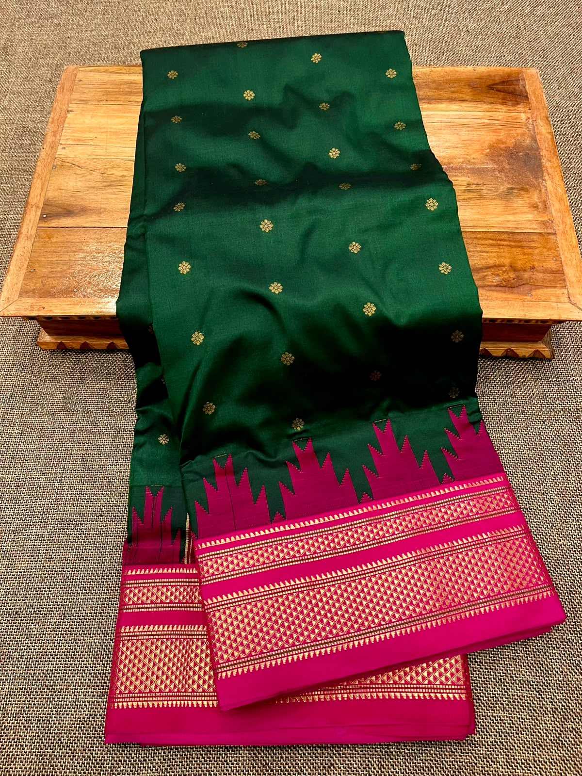 A dark green silk saree with small gold motifs and a vibrant magenta border, draped over a wooden tray on a textured gray carpet.