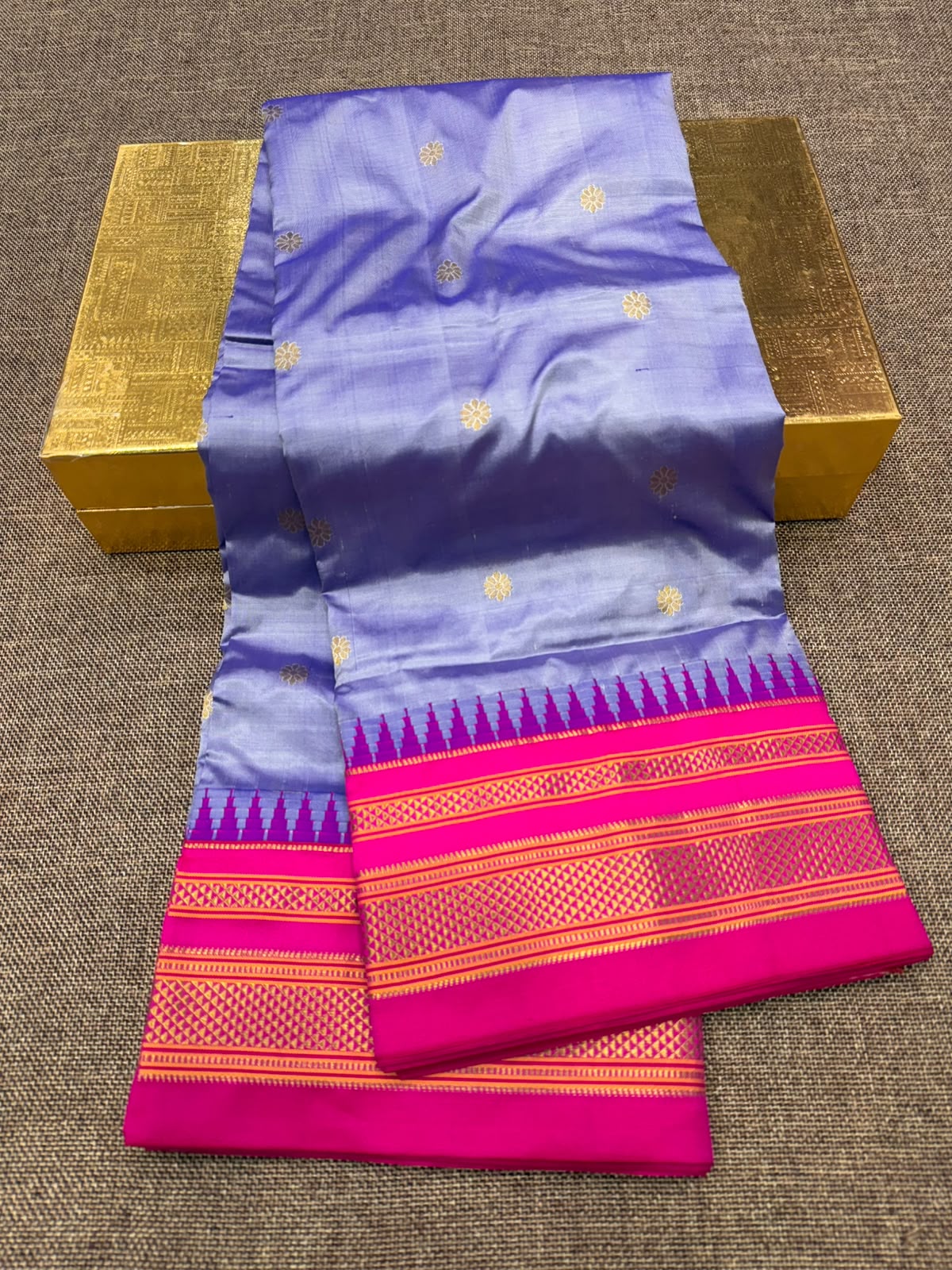 A folded purple silk saree with gold zari motifs and a vibrant magenta border, displayed on a textured gray surface with two gold boxes.