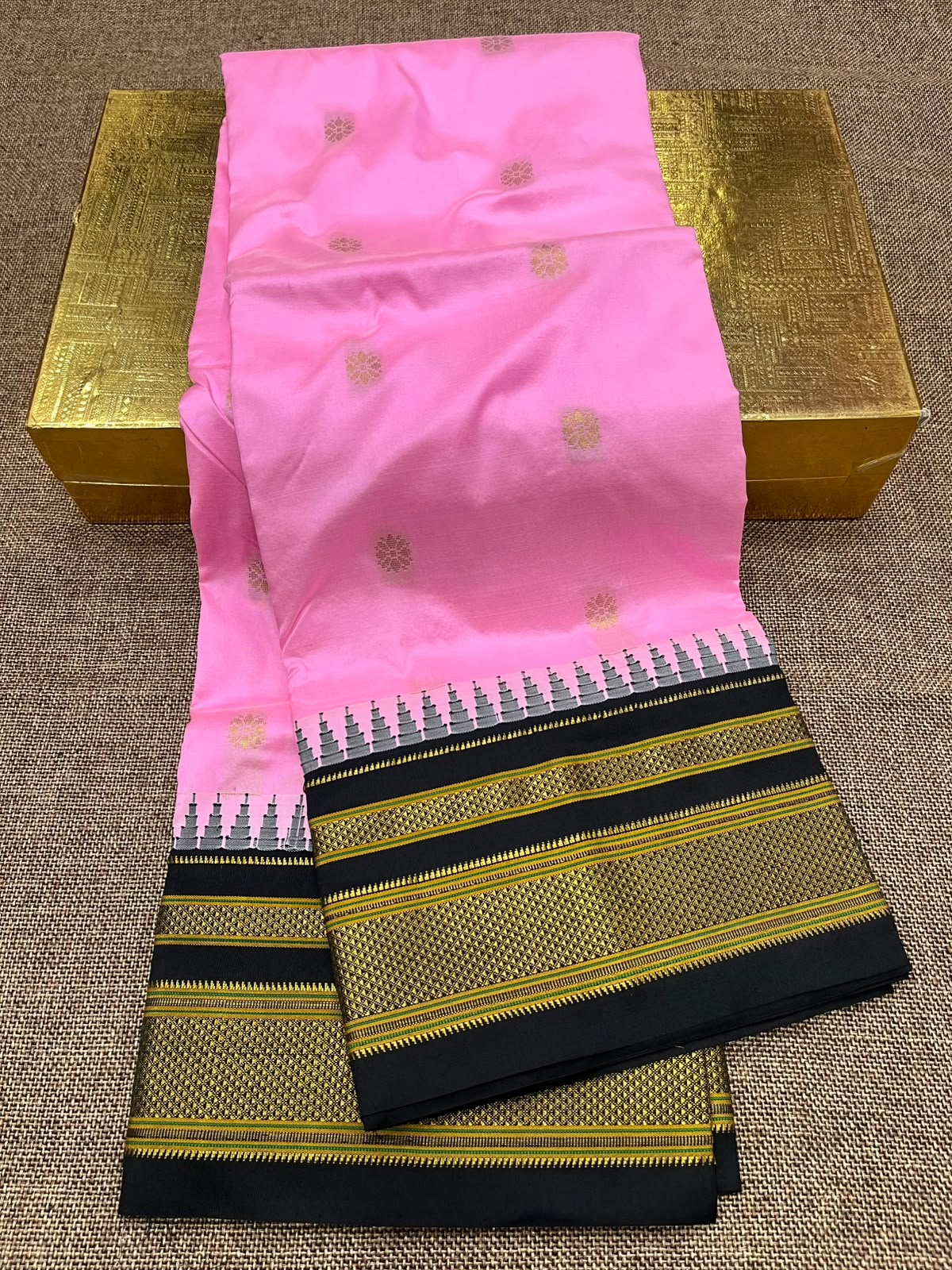 A folded pink saree with small gold motifs and a wide black, gold, and white zari border, displayed on a textured beige surface with a gold-colored box behind it.
