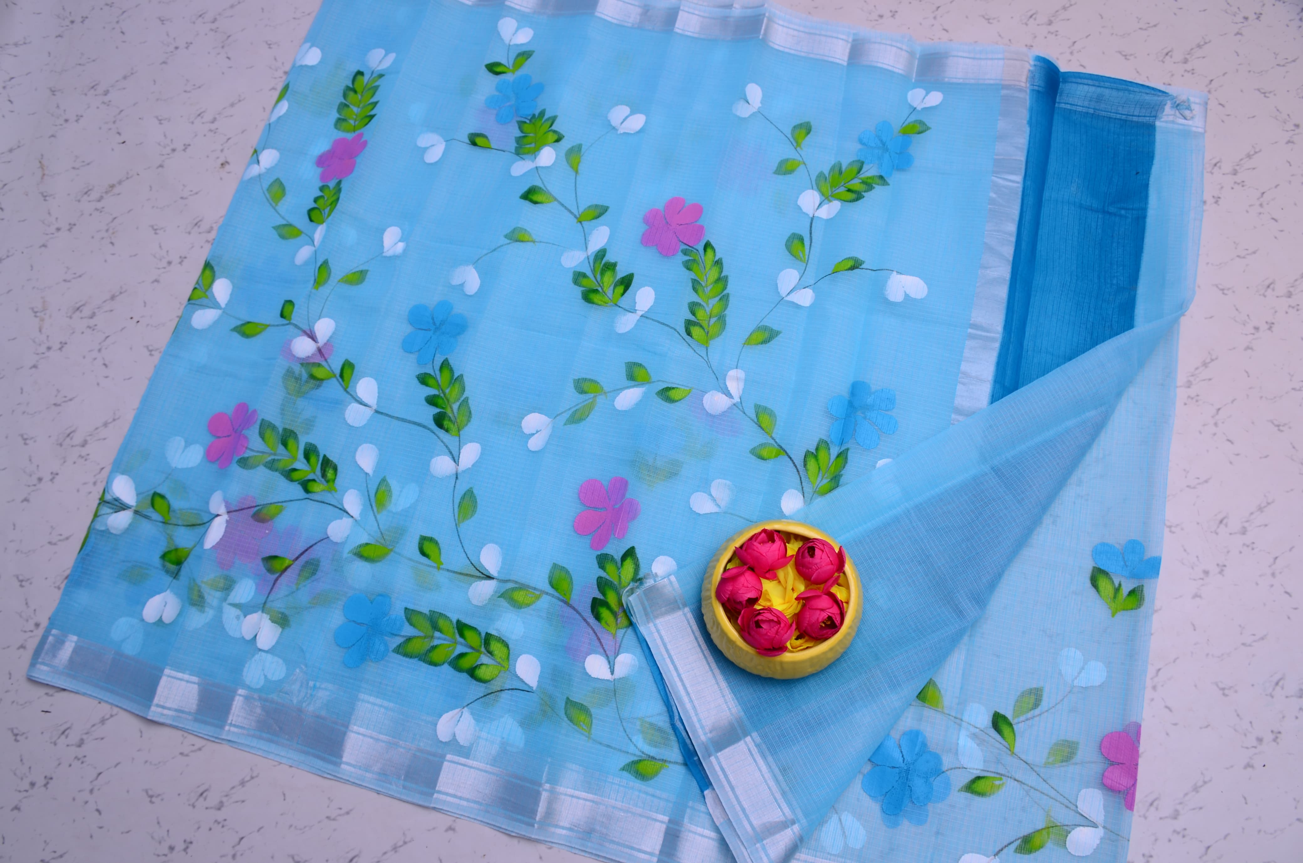 A light blue organza saree with floral embroidery of pink, blue, and white flowers with green leaves, laid flat on a light textured surface with a small yellow bowl of rose petals.