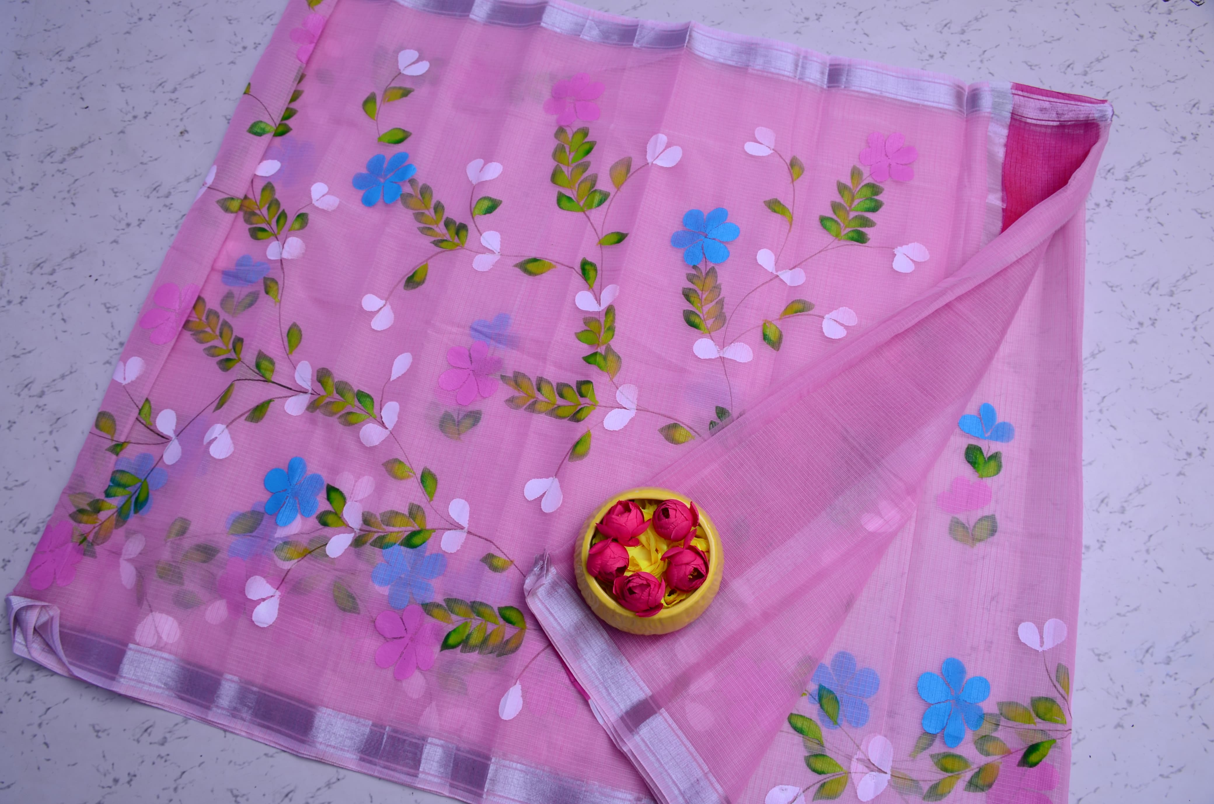 A pink sheer organza saree or scarf with embroidered floral patterns in blue, pink, and green, laid flat on a light gray textured surface with a small yellow bowl of rose petals in the center.