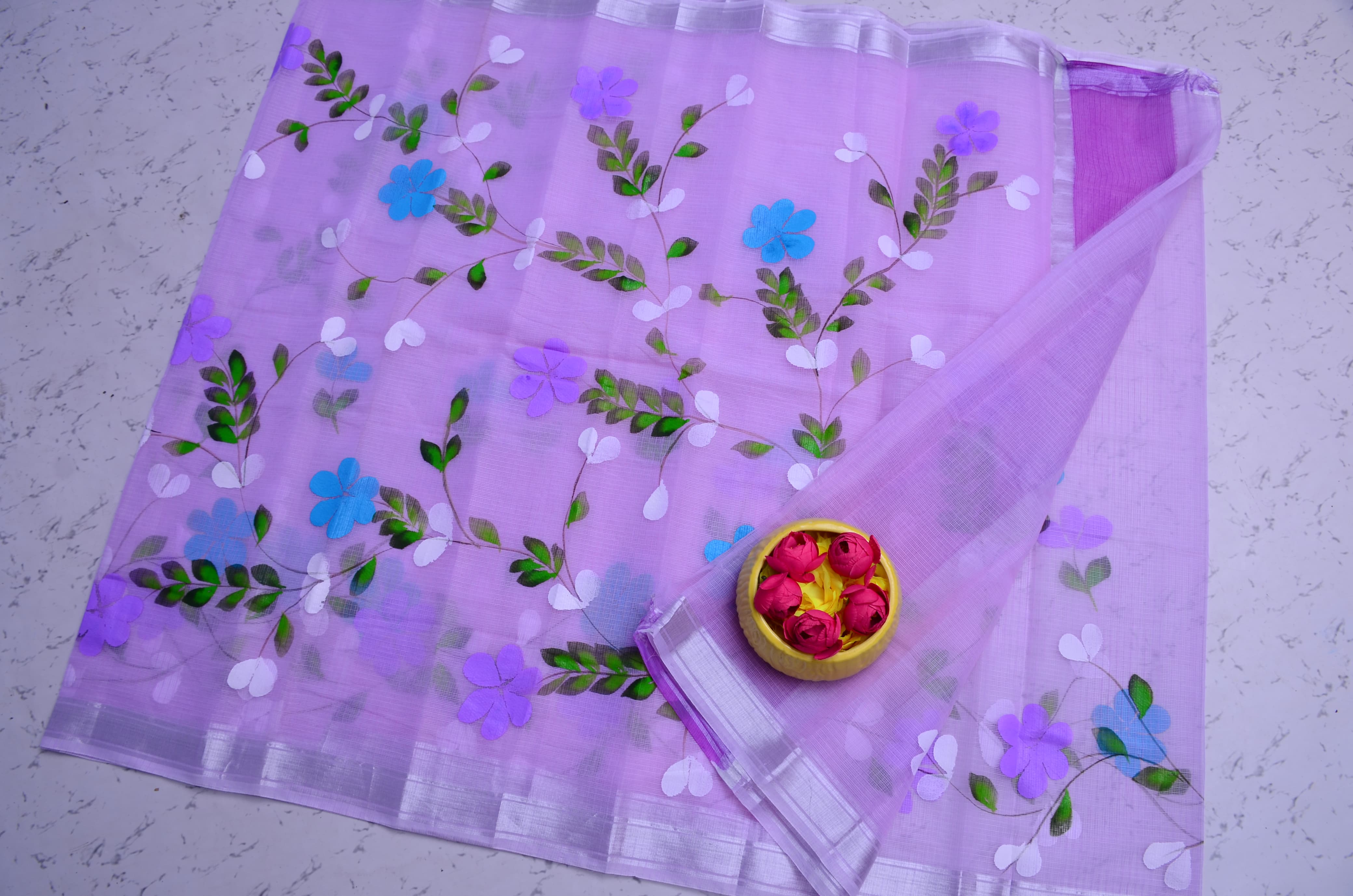 A lavender purple organza saree with embroidered floral patterns in blue, purple, green, and white, displayed on a light gray textured surface with a small decorative pot.