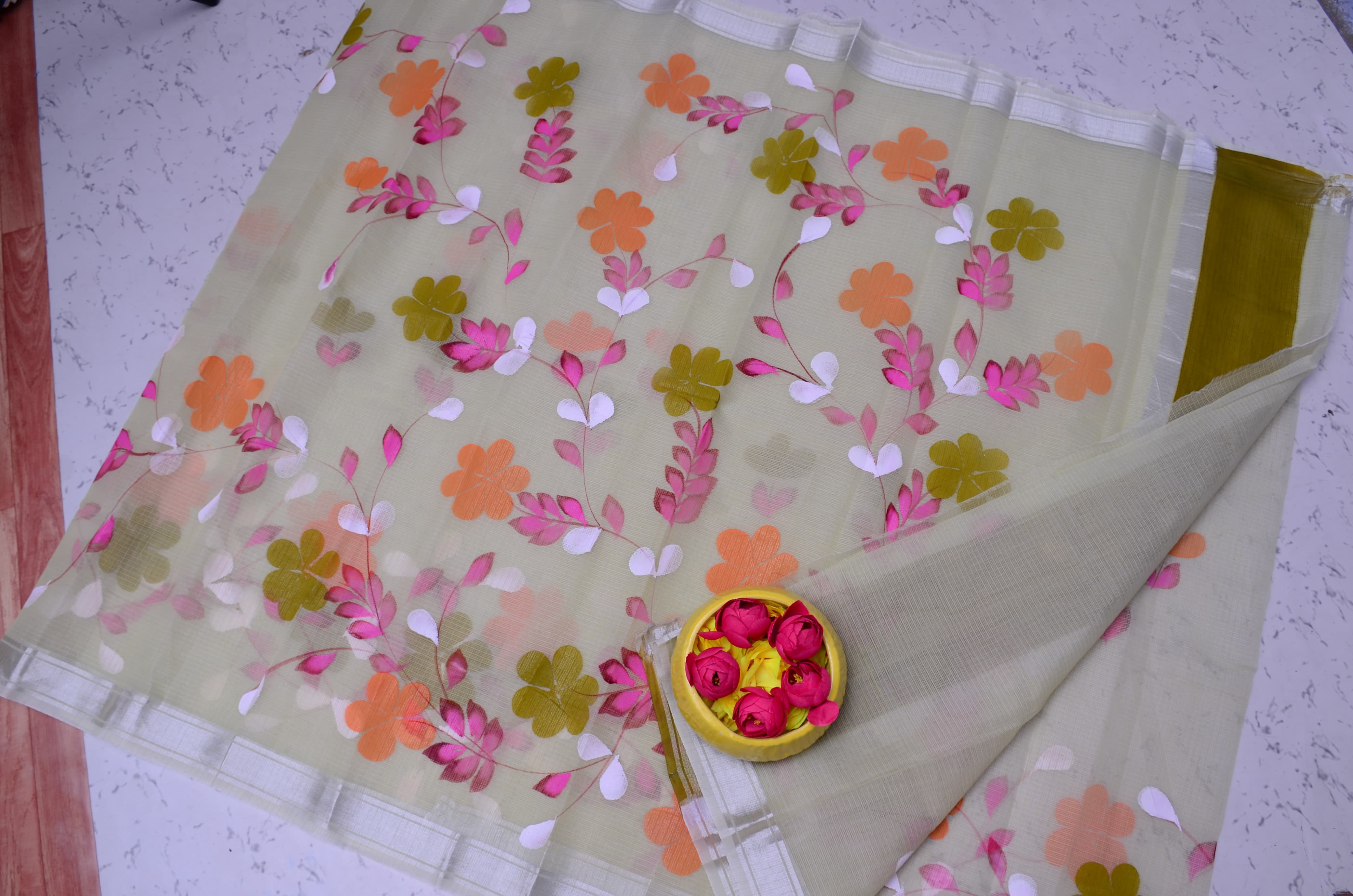 A semi-sheer beige saree or fabric piece with colorful floral embroidery in pink, orange, and olive green, laid flat on a speckled white surface with a small decorative bowl of pink flowers.