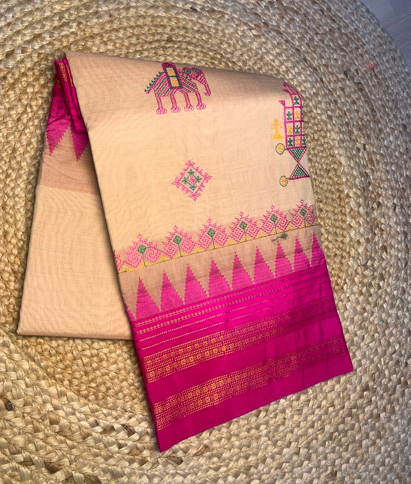 A folded beige and magenta Banarasi silk saree with traditional hand-embroidered motifs, displayed on a woven straw mat.