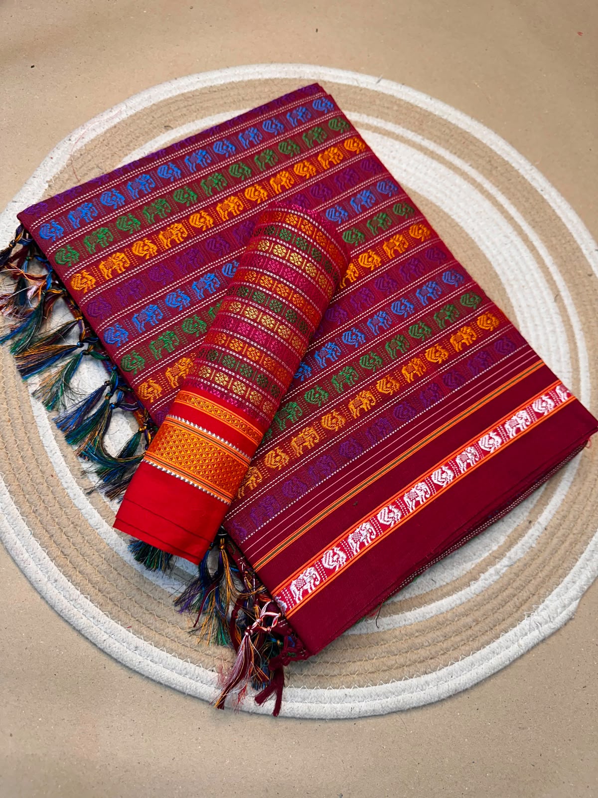 A folded and rolled multicolor patterned textile, likely a scarf, shawl, or sari, displayed on a round woven beige and white mat against a neutral beige background.