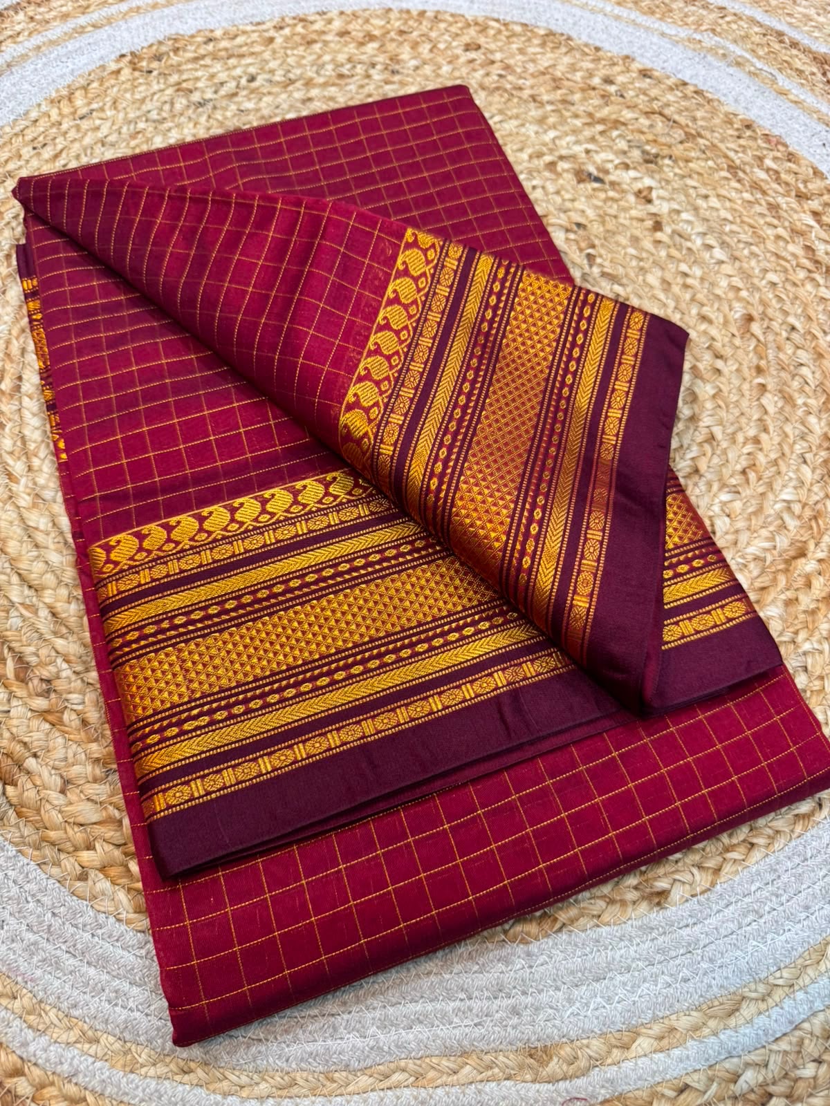 A folded traditional saree or fabric piece in deep maroon with yellow grid patterns and a wide golden-yellow border featuring intricate geometric and floral motifs, displayed on a woven straw mat.