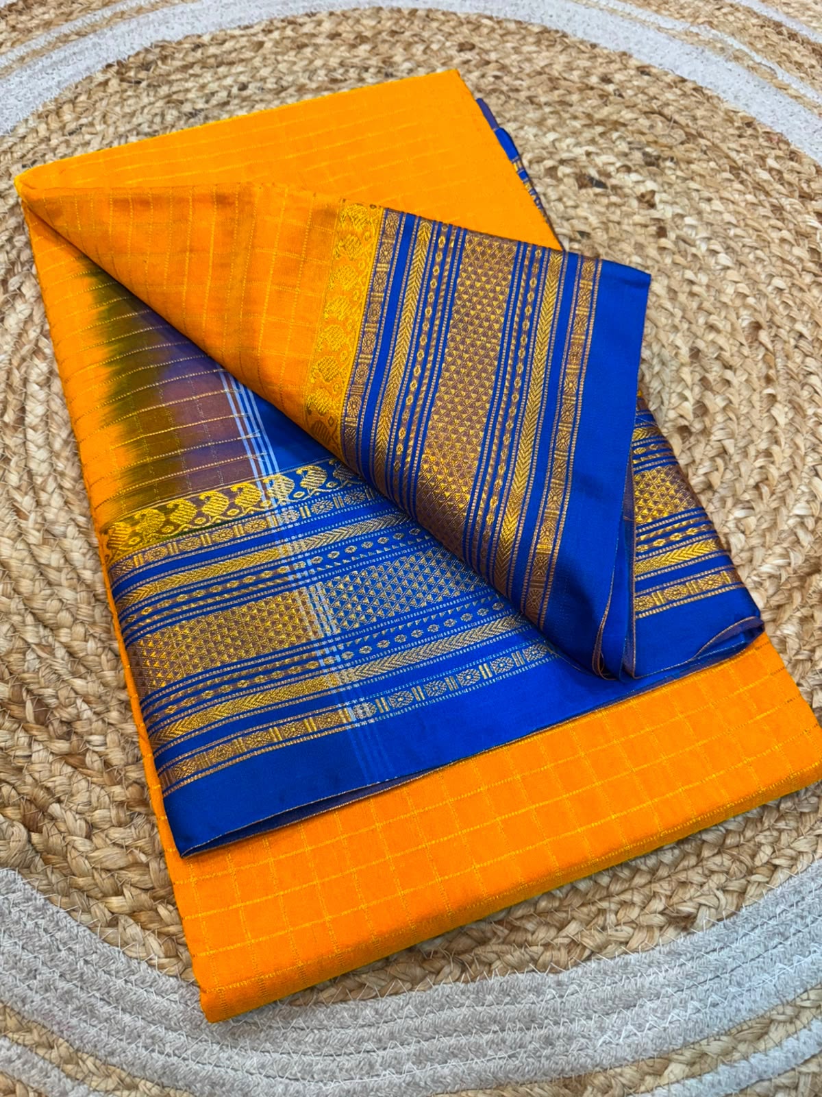 A folded orange and blue saree with intricate gold zari patterns, displayed on a round woven jute mat.