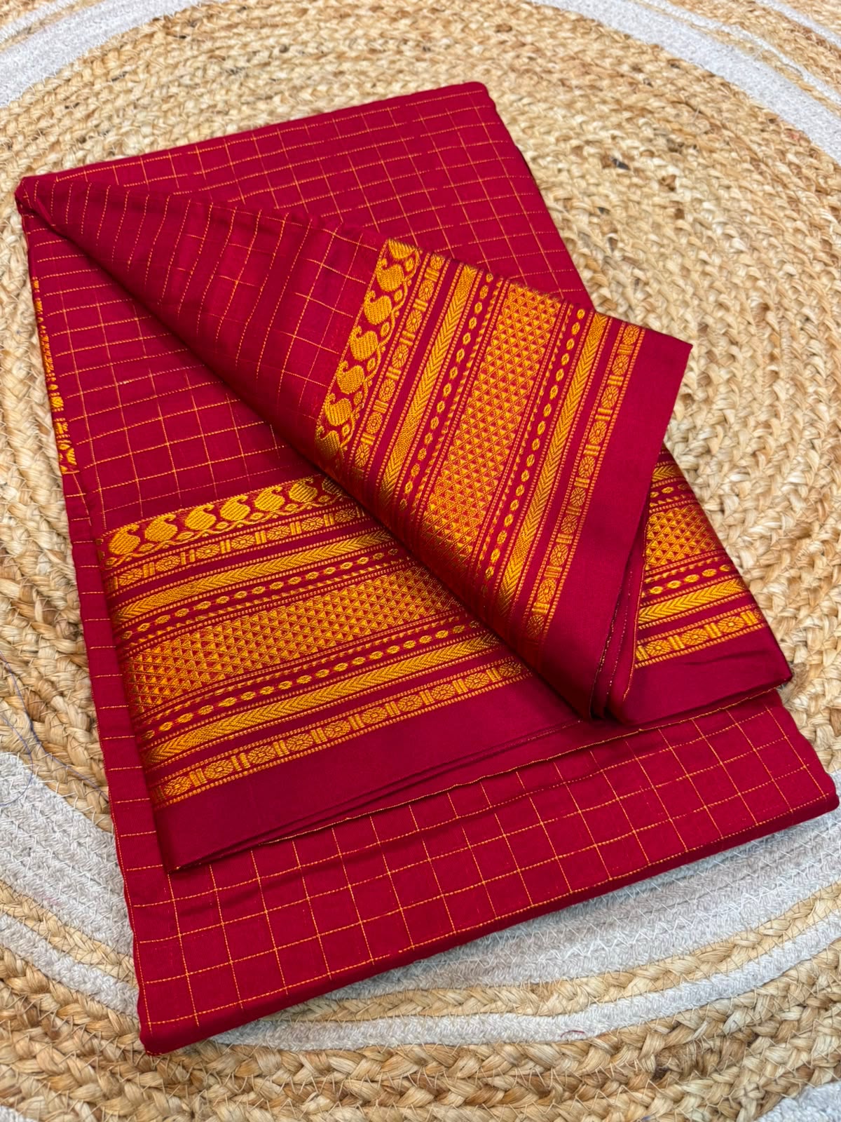 A folded red and orange patterned fabric, likely a saree or textile, displayed on a round woven jute mat. The fabric features a red base with fine yellow grid lines and a prominent orange and yellow traditional border design.