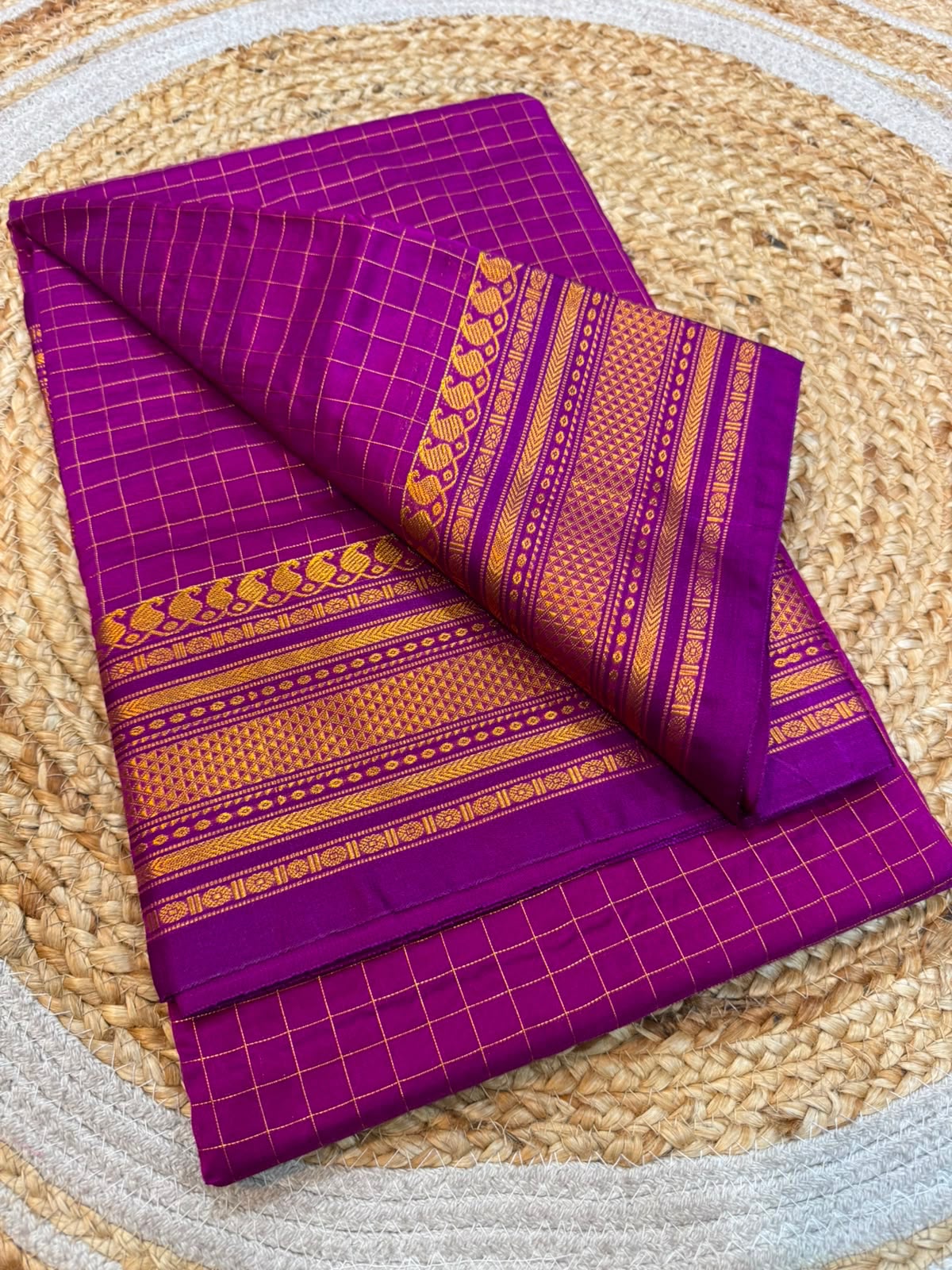 A folded purple saree with gold zari checkered grid patterns and ornate border designs, displayed on a round woven jute mat.