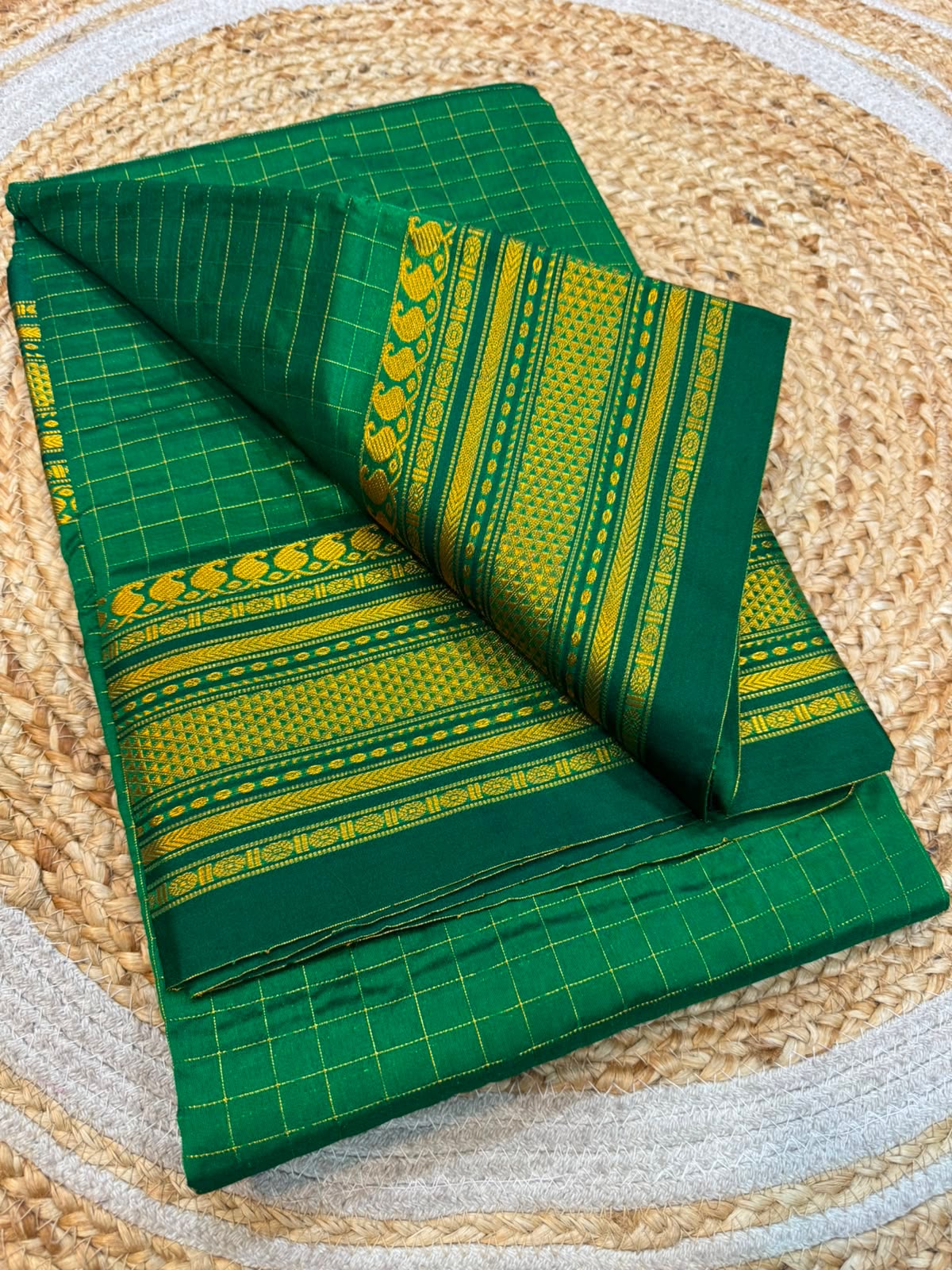 A folded green saree with yellow and green geometric patterns and borders, displayed on a round woven jute mat.
