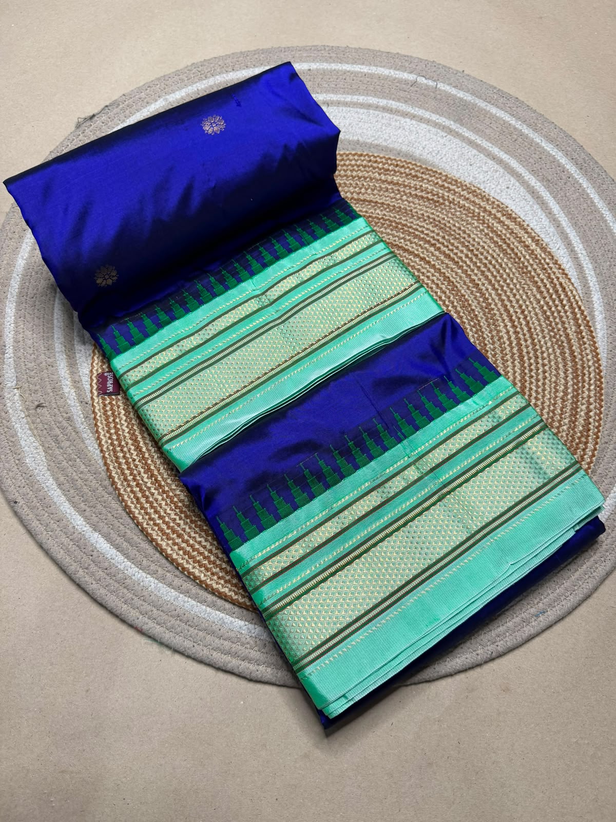 A folded royal blue and mint green saree with gold zari stripes, displayed on a circular woven placemat over a neutral surface.
