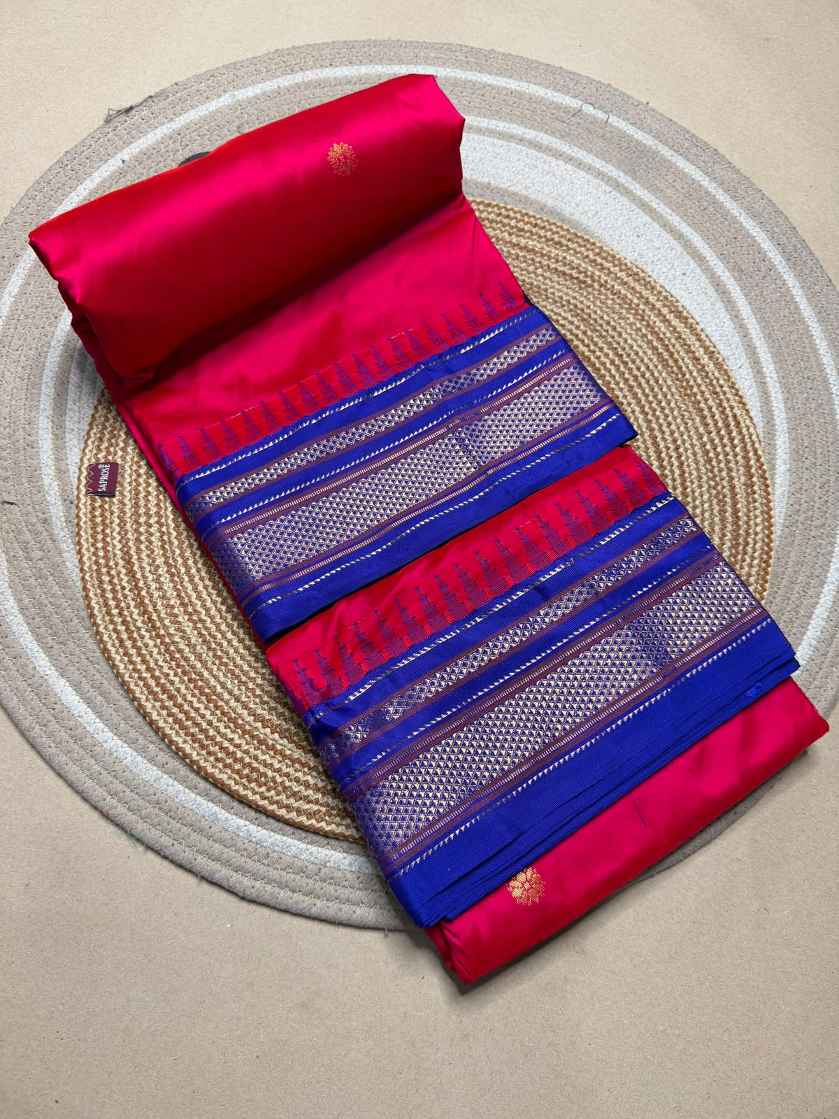 A folded red and blue saree with metallic zari borders, displayed diagonally on a round woven jute placemat over a neutral beige surface.