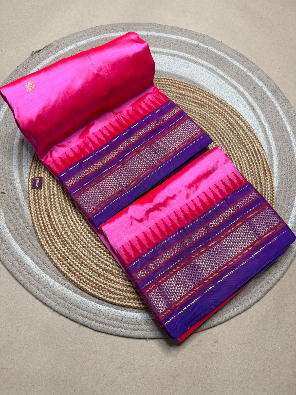 A folded and rolled pink and purple saree displayed on a round woven placemat over a neutral surface.