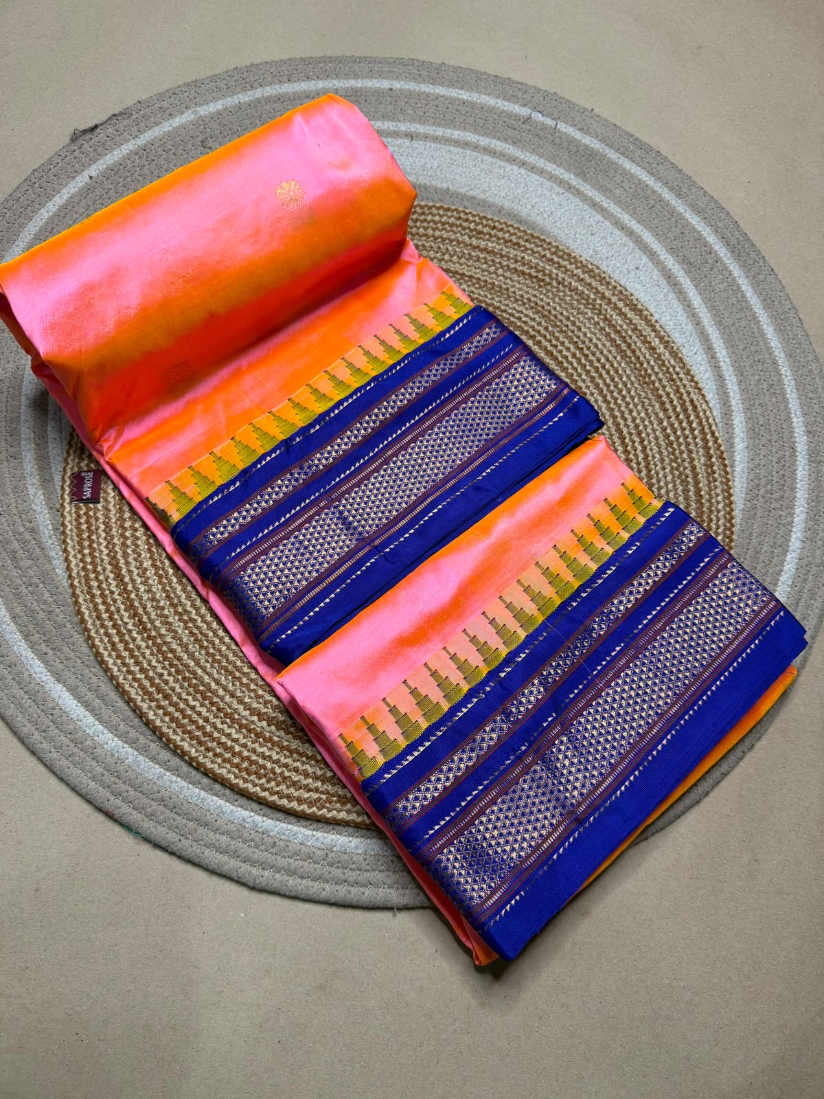 A folded and rolled saree displayed diagonally on a woven round mat. The fabric features a vibrant pink body with a bright orange border, decorated with multiple blue and purple borders containing gold and red zari details.