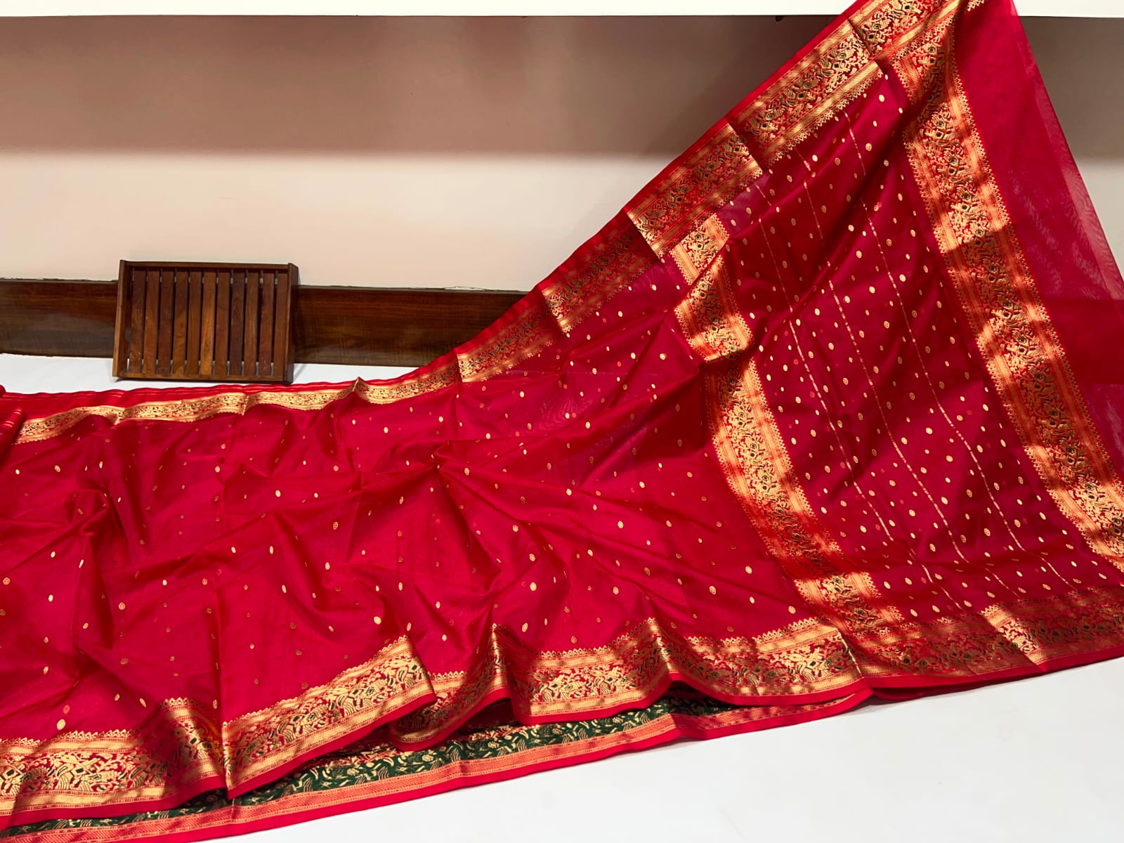 A red silk saree with intricate gold zari borders and small gold motifs, draped diagonally across a white surface.