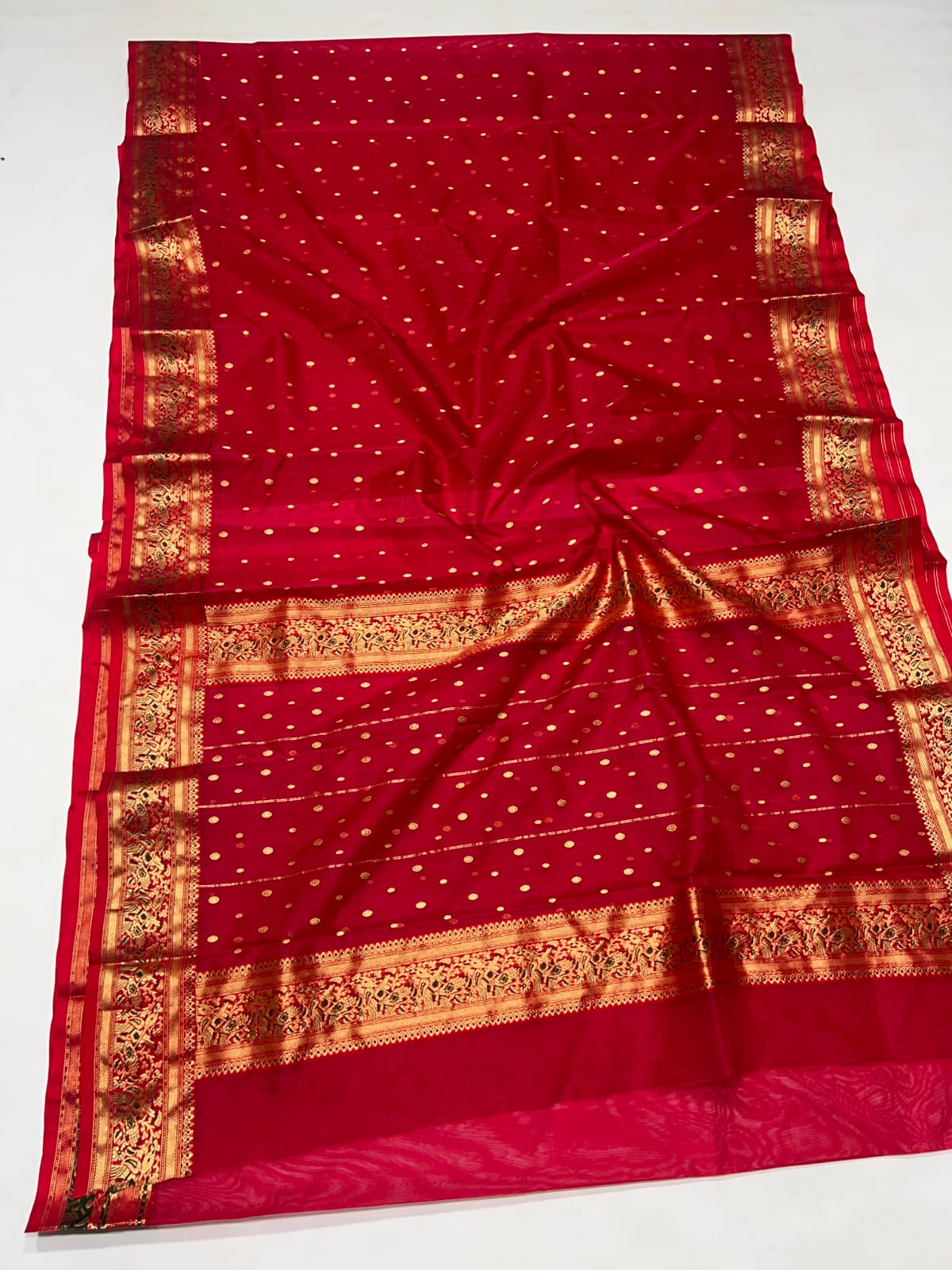 A deep red silk saree with small gold motifs and wide gold zari borders, laid flat on a white background.