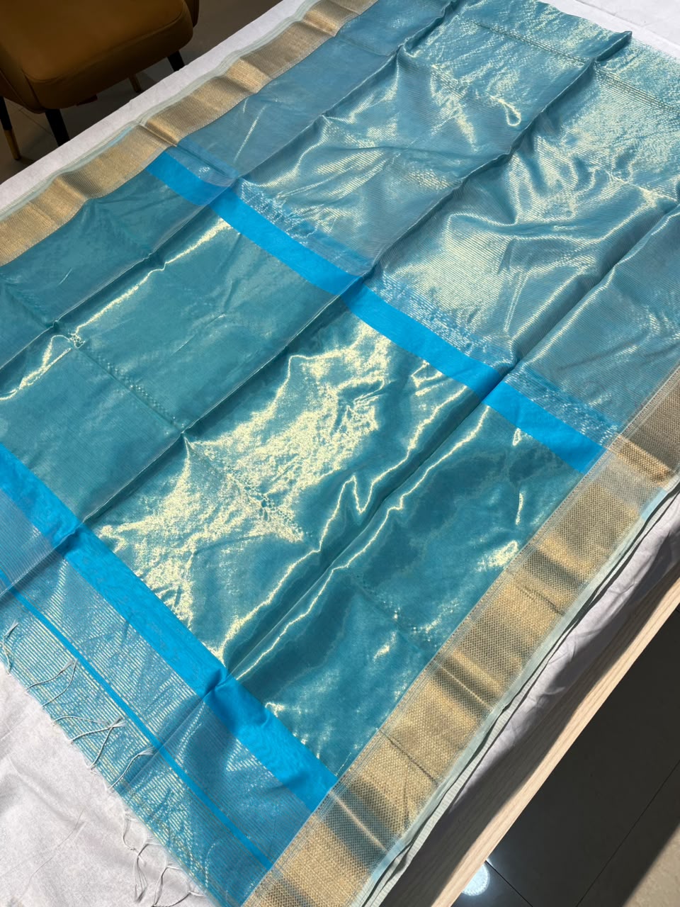 A bright turquoise blue silk saree with a metallic gold border, laid flat on a white surface.
