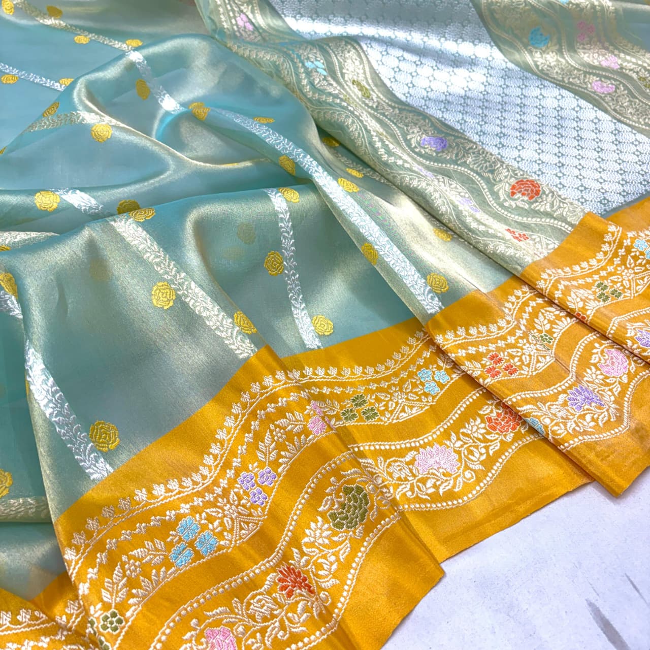 A close-up of a light blue-green sheer fabric with metallic silver stripes, gold floral motifs, and a wide mustard yellow border decorated with intricate multicolor embroidery.