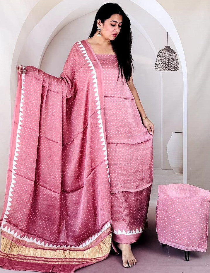 A woman models a pink silk salwar suit with white geometric borders and a gold zari border, paired with matching pink pants. She stands in a white arched corridor with a matching pink stool.
