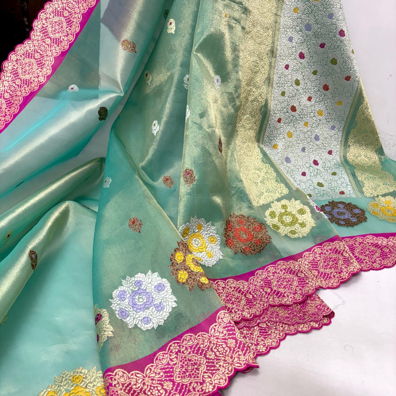 A close-up of a mint green silk saree or fabric with colorful floral embroidery, gold zari motifs, and a bright pink border with intricate gold woven detailing.