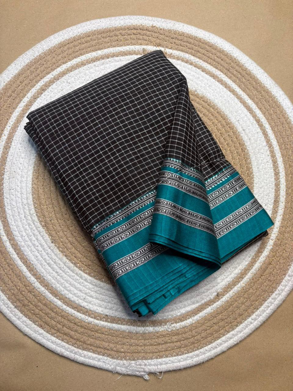 A folded dark gray and white checkered fabric saree with a teal border, displayed on a circular woven mat over a tan surface.