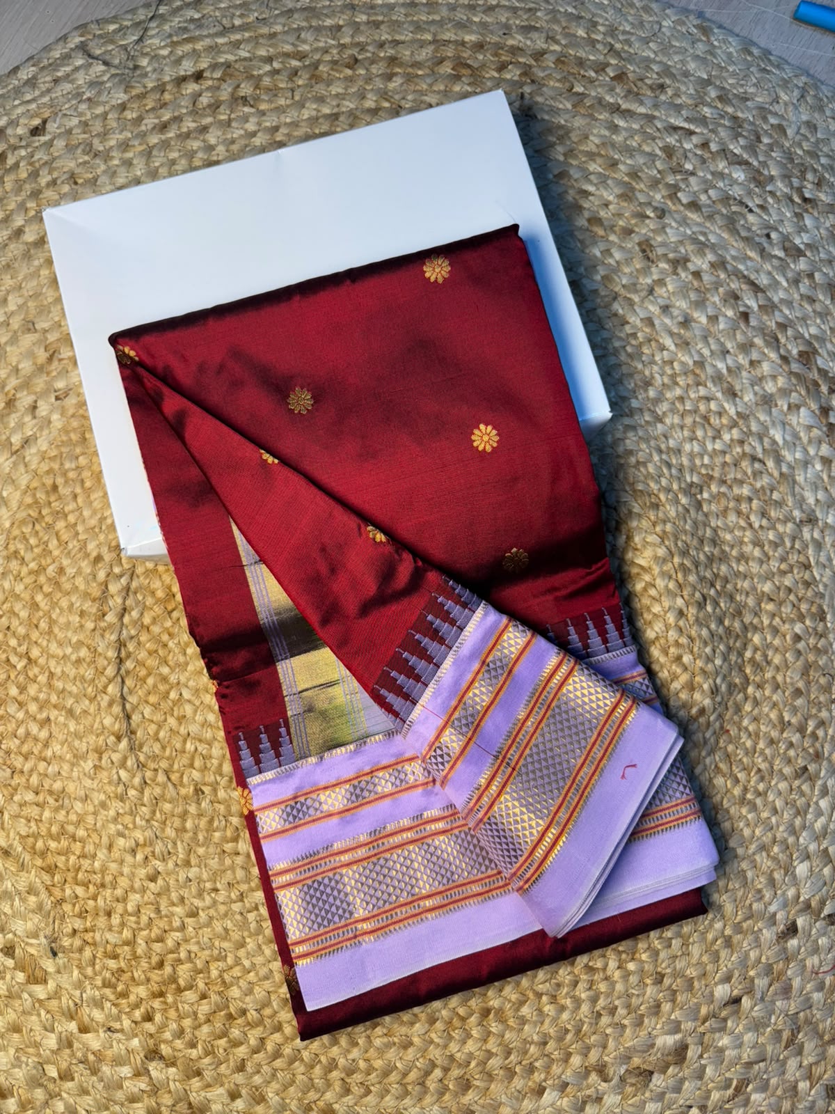 A folded red and lavender saree with gold zari motifs and a patterned border, displayed on a white square card over a woven straw mat.