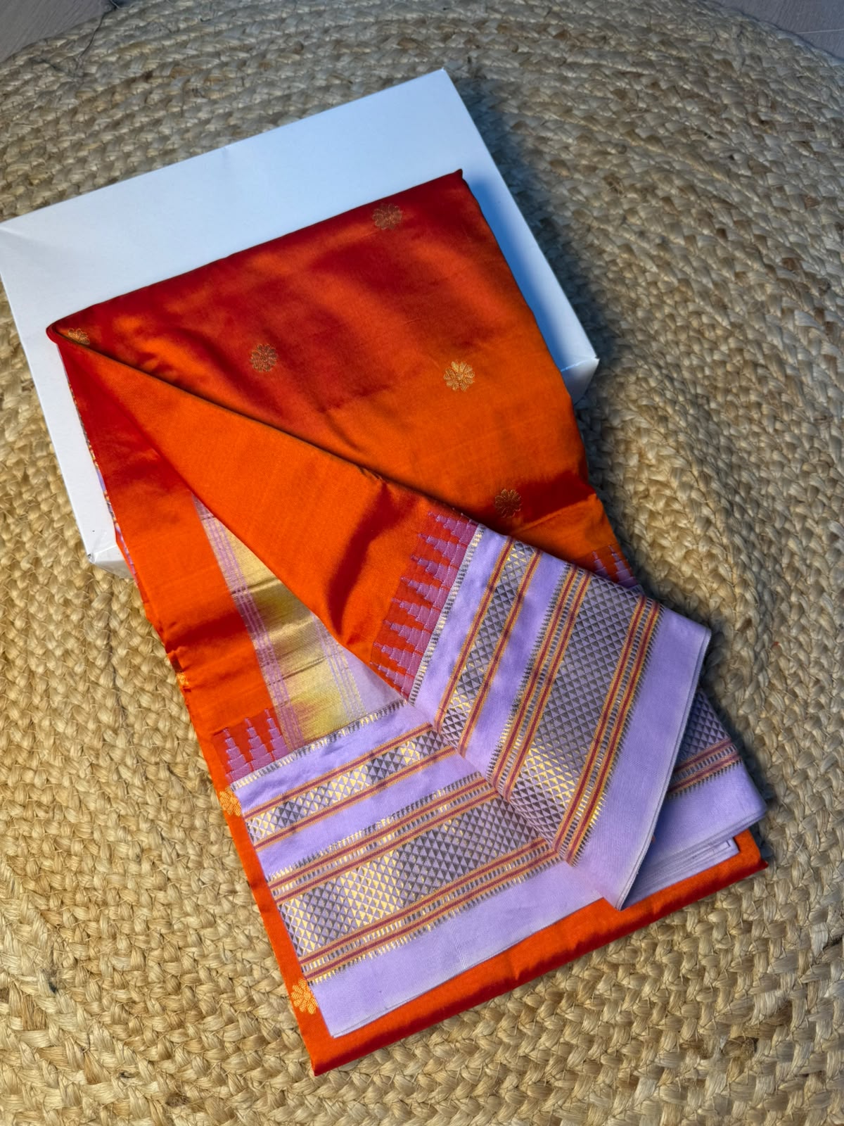 A folded silk saree with a vibrant orange-red body and a lavender-purple border with gold zari detailing, displayed on a white box over a woven jute mat.