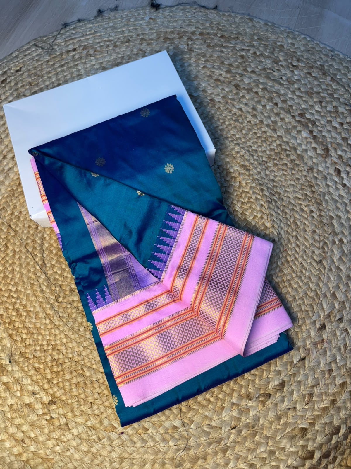 A folded silk saree with a deep teal blue body, pink border, and gold zari detailing, displayed on a woven straw mat next to a white box.